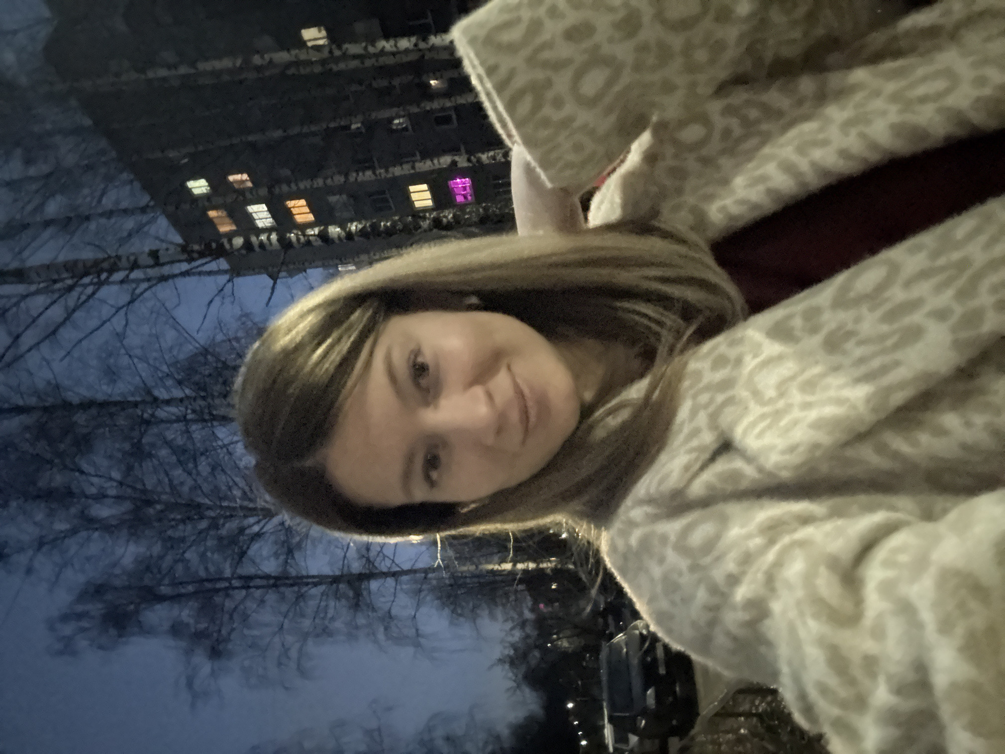 A woman is taking a selfie at night. In the background, there is a multi-story building with several illuminated windows, including one with a pink light. The trees are bare, and the overall lighting suggests it is dusk or night. The location is plausibly Chaikovsky, Russia, based on the architectural style of the building.