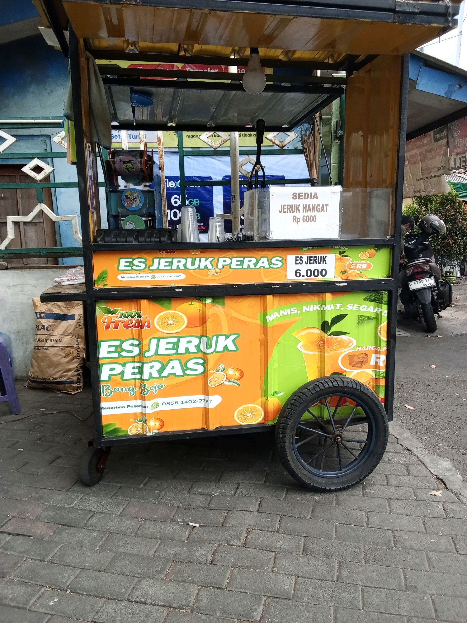This is a medium shot of an orange juice cart positioned on a brick sidewalk. The cart is black and yellow with a large orange and green advertisement panel. The panel displays images of oranges and text in Indonesian, advertising "ES JERUK PERAS" (freshly squeezed orange juice). One section of the advertisement indicates "ES JERUK Rp 6.000" (orange juice Rp 6,000). Another section reads "MANIS, NIKMAT, SEGAR L" (sweet, delicious, fresh). There is a black bicycle wheel attached to the right side of the cart. To the left of the cart, there is a brown paper bag with text in Indonesian and English, including "MALAYSIA". In the background, a motorcycle with a helmet on its seat is parked. The environment suggests an outdoor urban setting, possibly a street in South Jakarta, Indonesia, given the presence of Indonesian text and a common street vending setup.