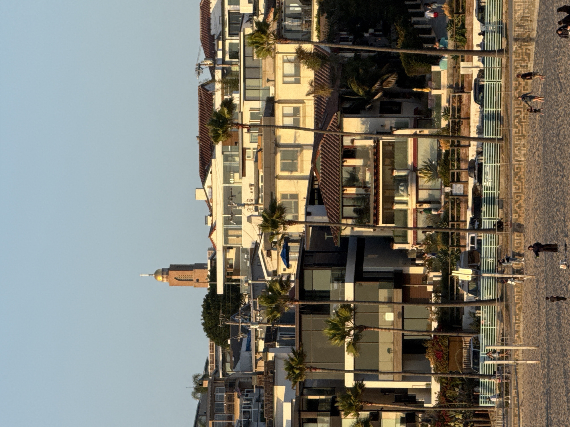 The image depicts a beach scene in Manhattan Beach, California, United States of America. In the foreground, a sandy beach with scattered people is visible. A volleyball net setup is present. In the midground, a light green horizontal fence separates the beach from a street lined with parked cars and a few people walking. Beyond the street, a row of modern beach houses with palm trees in front are visible. In the background, a tall brick tower with a golden dome and a white spire stands out against the clear blue sky, a prominent architectural feature in the city. The overall lighting suggests it is late afternoon or early evening due to the warm sunlight casting shadows.