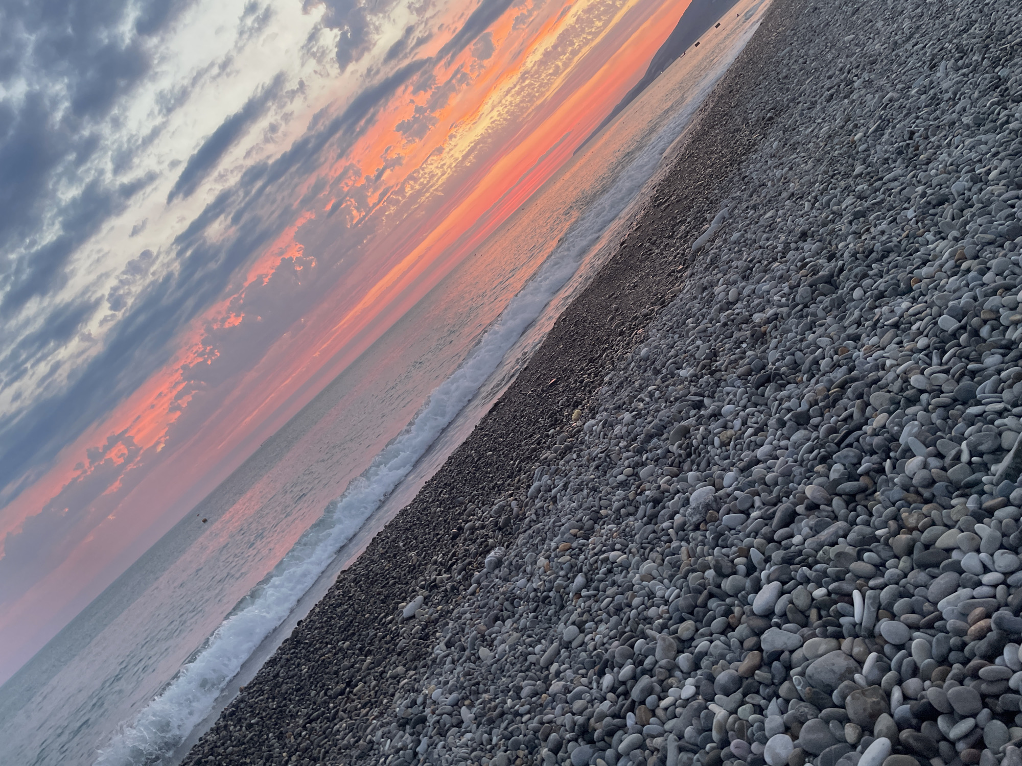 A diagonal shot depicts a pebble beach meeting the ocean at sunset, with a striking pink and orange sky. The foreground is filled with smooth, grey pebbles of varying sizes, some wet from the receding tide. A gentle wave breaks along the shoreline, its white foam contrasting with the darker, wet pebbles. The ocean stretches towards the horizon, reflecting the vibrant colors of the sunset. Wispy clouds in shades of grey, pink, and orange stretch across the sky, creating a dramatic backdrop. The image is oriented diagonally, giving a sense of perspective and depth. The scene is devoid of people or any man-made structures, emphasizing the natural beauty of the environment. The overall mood is serene and tranquil, capturing the fleeting moment of twilight.
