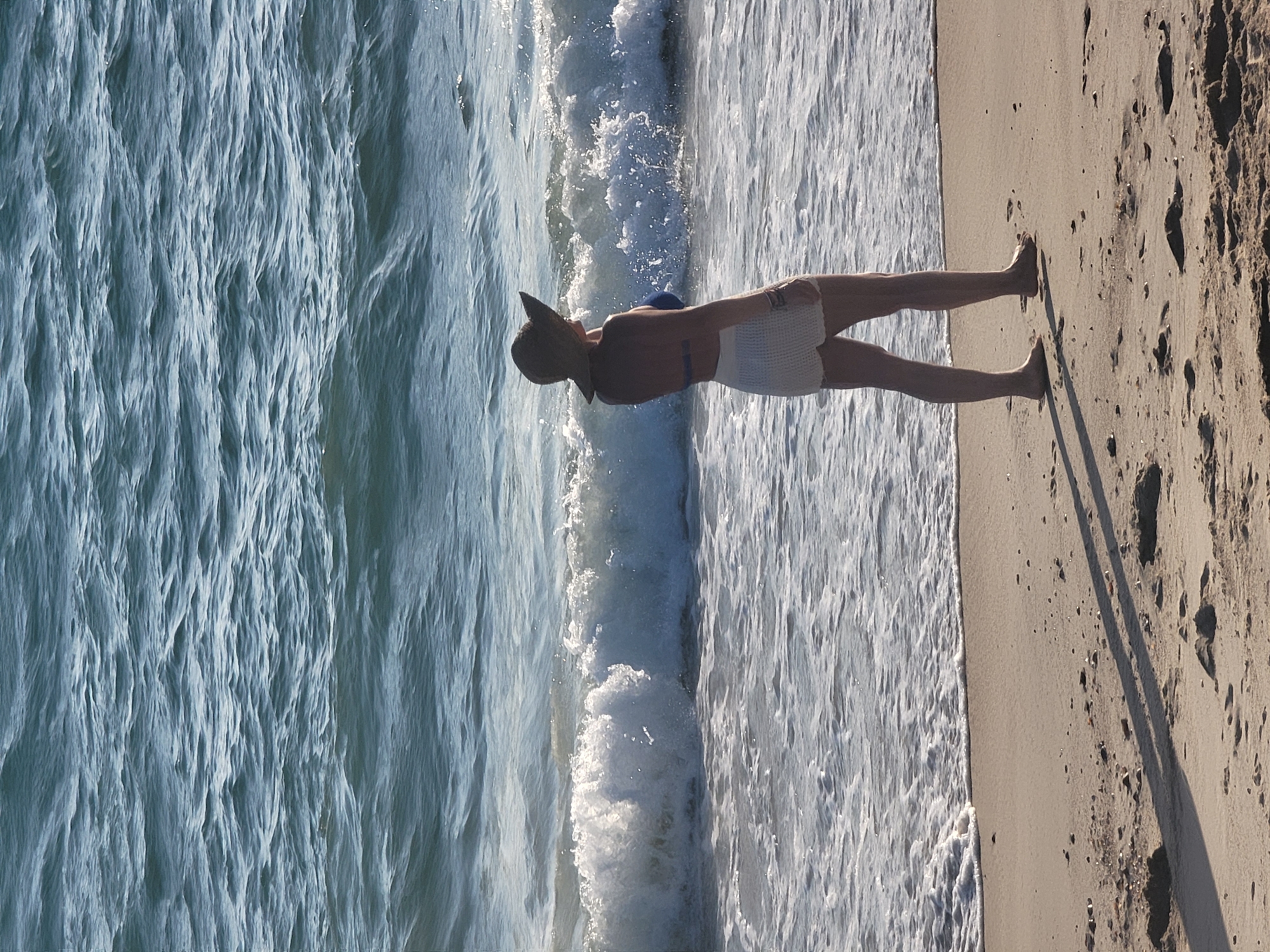 A person wearing a wide-brimmed hat and a white crocheted skirt over a blue bikini stands on the sandy shore of Quarteira, Portugal, facing the ocean. Waves are breaking nearby, creating a foamy white surf that washes onto the beach. The sunlight casts a shadow of the person onto the sand. The ocean is a deep blue with visible ripples, and the sky is a pale blue. The scene appears to be a serene beach setting.
