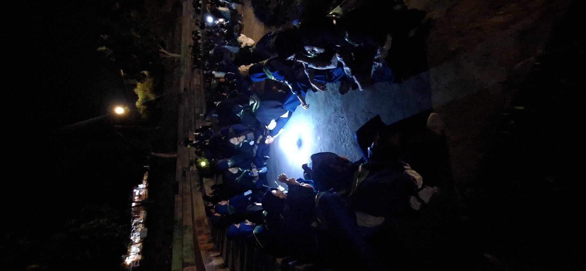 In Karang, Indonesia, at night, a group of people are gathered in an outdoor amphitheater. They are sitting in a circle on the ground, illuminated by a bright light emanating from the center. Many of them are wearing dark clothing with blue accents and some have green scarves. Some individuals are holding what appear to be small lights, possibly flashlights or phone screens. In the background, there are trees and a street with shops dimly lit. The overall atmosphere suggests a communal gathering or activity taking place after dark.