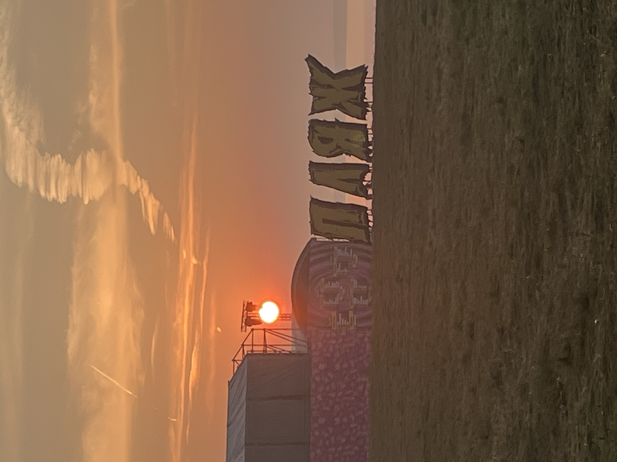 The image depicts a festival or event setup at dusk in Surakovo, Russia. In the background, the setting sun illuminates the sky with warm orange and yellow hues. A stage or tent structure is visible on the left, with lights mounted on scaffolding. In front of this structure, large, stylized letters spelling "ПЛЯЖ" (meaning "BEACH" in Russian) are prominently displayed. The letters are positioned on a grassy field, with the sea or a large body of water visible in the distance behind them. The overall scene suggests an outdoor event taking place by the coast during a sunset.