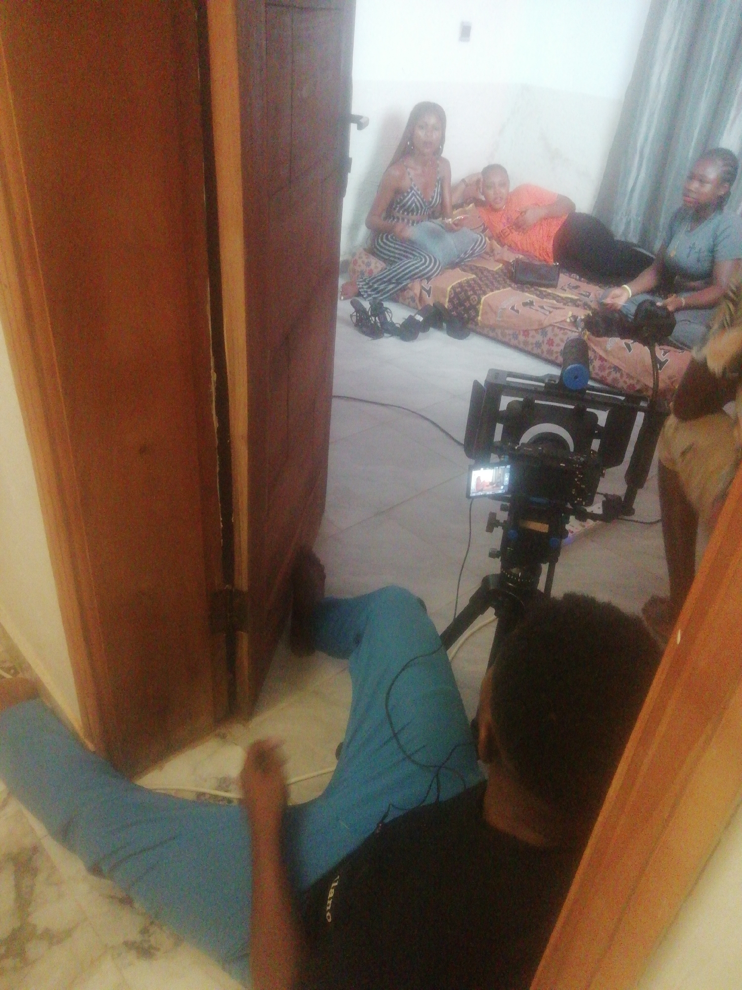 The image depicts an indoor video production scene, likely in Ikorodu, Nigeria. In the foreground, a person, presumably the cameraman, is seated on the tiled floor, wearing blue pants and a black shirt with "tano" visible on it. They are operating a professional-grade video camera mounted on a tripod. The camera rig includes a small monitor displaying an image, and what appears to be a shotgun microphone.
The cameraman is positioned through an open, tall wooden door, which frames the subjects of the shoot. Beyond the door, three women are casually seated and reclining on a patterned mattress or floor cushion. The woman on the left, wearing a black and white striped jumpsuit, looks towards the camera while holding a small item. The woman in the center, dressed in an orange t-shirt, reclines comfortably, also facing the camera. To her right, a third woman in a light blue t-shirt is seated, engaged in conversation or observing the setup. Several pairs of shoes are scattered near the mattress. The background features light-colored walls and what might be a curtain on the right. The scene suggests a relaxed yet focused filming environment.
