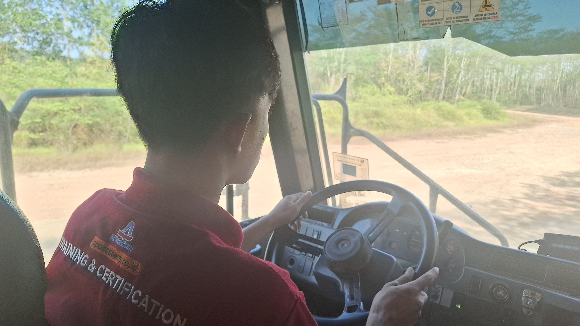 The image, captured from the interior of a vehicle, shows the back of a person's head and shoulders as they operate the steering wheel. The individual is wearing a red polo shirt with a logo and text that reads "TRAINING & CERTIFICATION" and "ppakdemy.co.id". The vehicle's dashboard is visible, featuring a steering wheel, various gauges, and a dashboard camera. Sunlight streams through the windshield, illuminating the dusty, unpaved road and sparse vegetation outside. The scene suggests an outdoor training or certification environment, possibly in a rural or industrial area of Indonesia, given the text on the shirt.