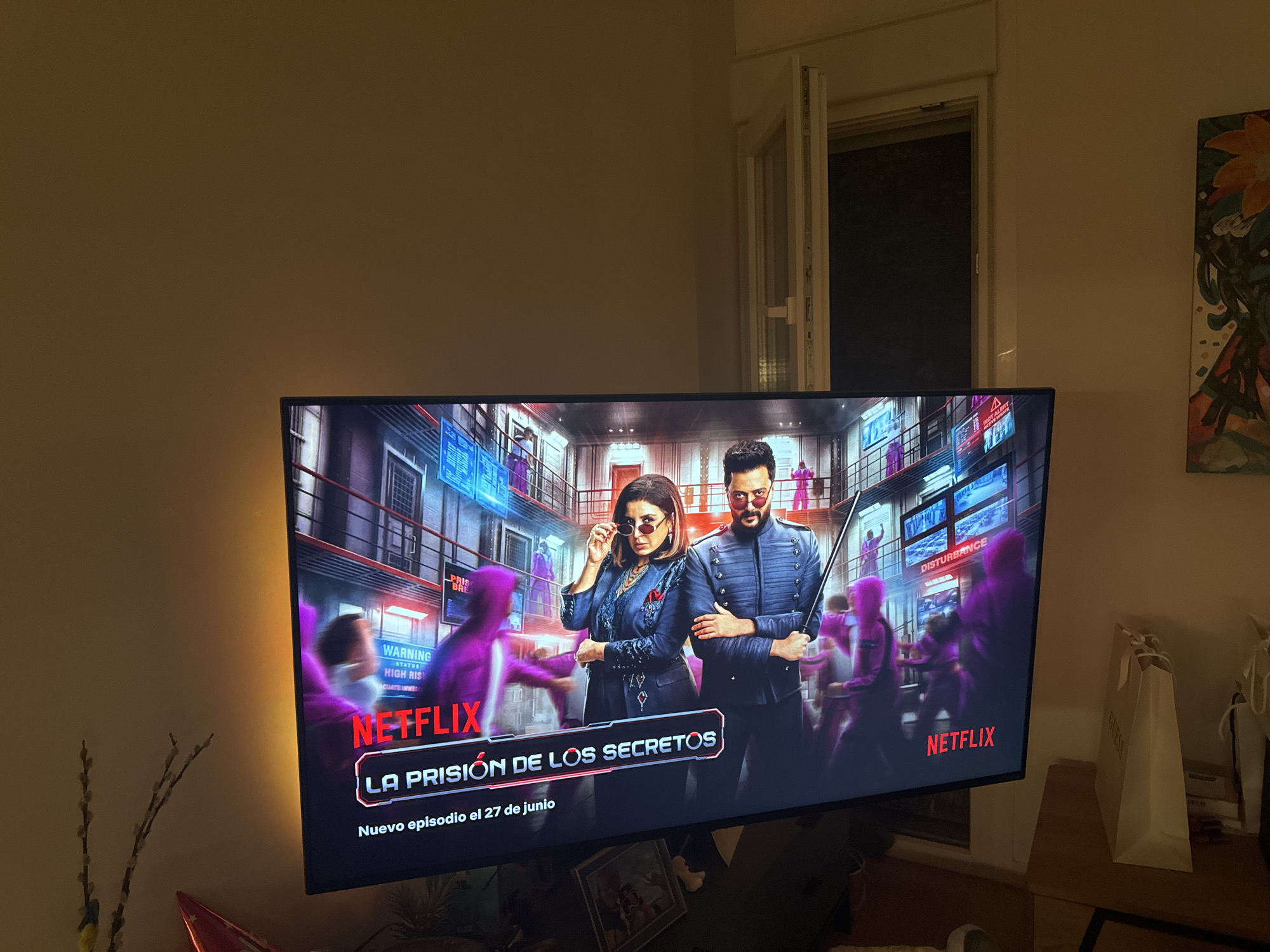 The image shows a television screen displaying a Netflix program titled "La Prisión de Los Secretos" (The Prison of Secrets). The screen features two main characters, a woman and a man, dressed in fashionable attire. The woman is wearing sunglasses and a blue jacket, while the man is in a blue military-style jacket and sunglasses, holding a sword. Behind them, figures in purple are blurred, suggesting a prison or a similar setting with multiple levels and screens displaying information. The Netflix logo is prominently displayed in red. The television is placed in a dimly lit room, with a warm yellow light emanating from the side of the screen. A window with an open pane is visible in the background, with darkness visible through it. To the right of the television, there is a painting of a flower on the wall and a light-colored shopping bag. The overall impression is a domestic setting where someone is watching a streaming service. There are no elements in the image that indicate a specific location in the city of Murcia, Spain.