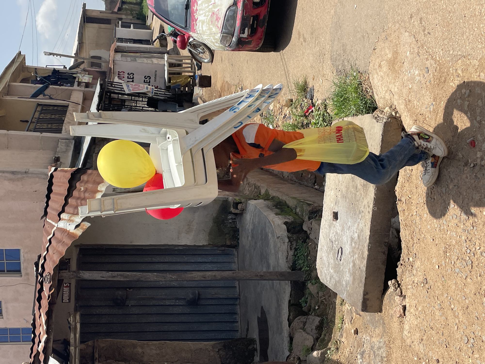 In the city of Jos, Nigeria, a young boy carries a stack of four white plastic chairs on his head, balanced precariously. One yellow balloon and one red balloon are tied to the topmost chair. He is wearing an orange t-shirt, blue jeans, and white sneakers. In his right hand, he holds a yellow plastic bag with the word "THANK" printed in red. The boy is walking on a dirt road, with a concrete slab to his left and a building with a corrugated metal roof to his left. A red car is parked to his right, and a motorcycle is visible behind it. The environment appears to be a residential or commercial area with various buildings and some overgrown vegetation.