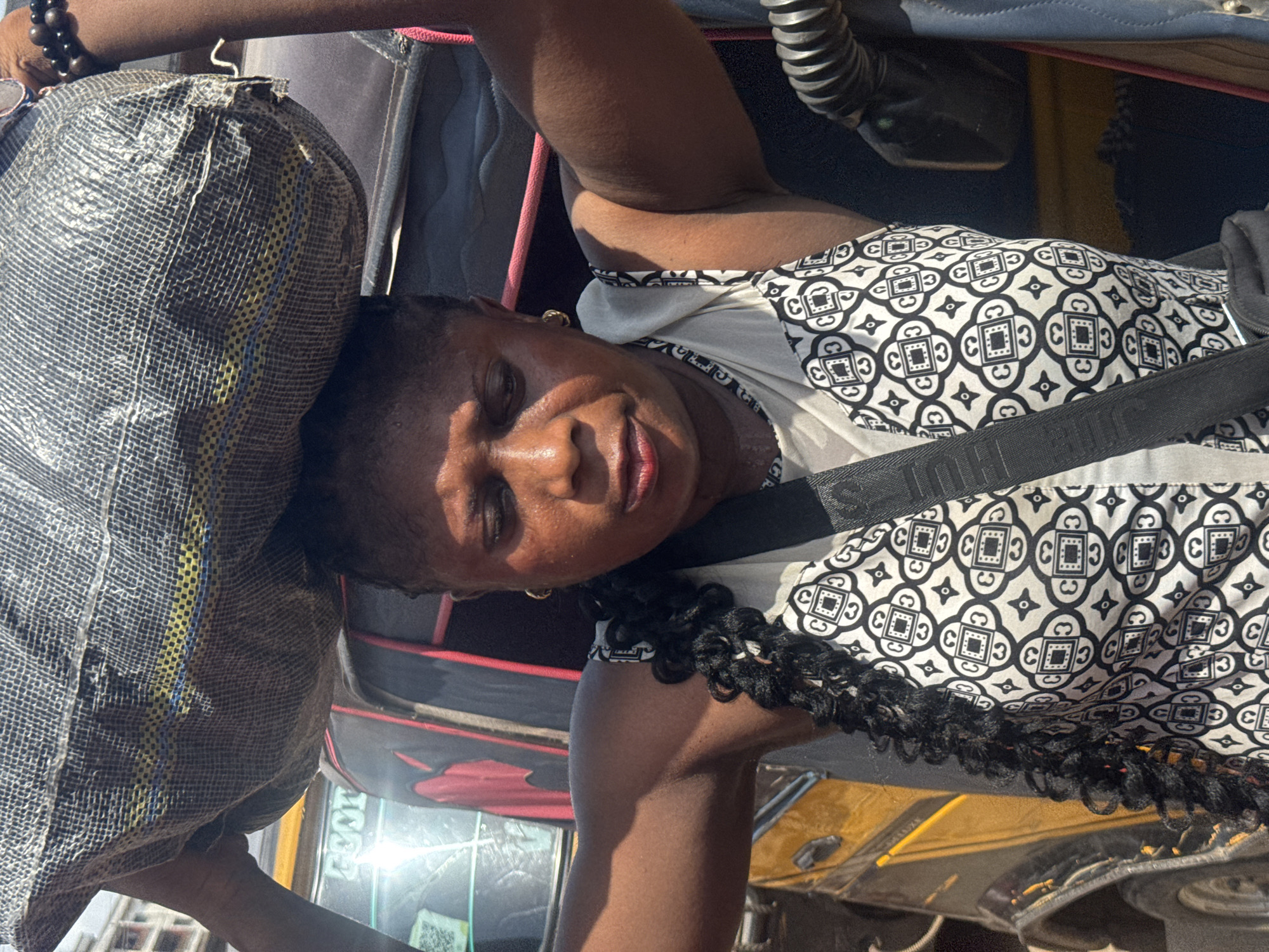 An image, rotated 90 degrees, depicts a person, likely a woman, seated in what appears to be a vehicle in Onitsha, Nigeria. She has dark skin and is looking towards the camera with a relaxed expression, her face illuminated by bright daylight, suggesting a sunny daytime setting.
She is wearing a sleeveless top featuring a distinctive black and white geometric pattern. A black strap, likely a seatbelt or a bag strap, crosses her chest diagonally. Her hair is styled in dark braids or twists, with some strands visible over her shoulder. A small gold earring is visible on her left ear, and a beaded bracelet adorns her right wrist.
A large, gray, woven synthetic bag with prominent yellow and blue stripes rests near her, suggesting she is traveling with luggage. The background elements indicate an enclosed space, consistent with a vehicle interior, showing upholstery and reflections through what may be a window.
Visible text from OCR includes "GEOT" on the black strap, possibly part of a brand name. Other detected characters ("C", "3", "O", "బి", "CC", "36", "ε") are largely interpretations of the geometric patterns on her clothing and not legible words.