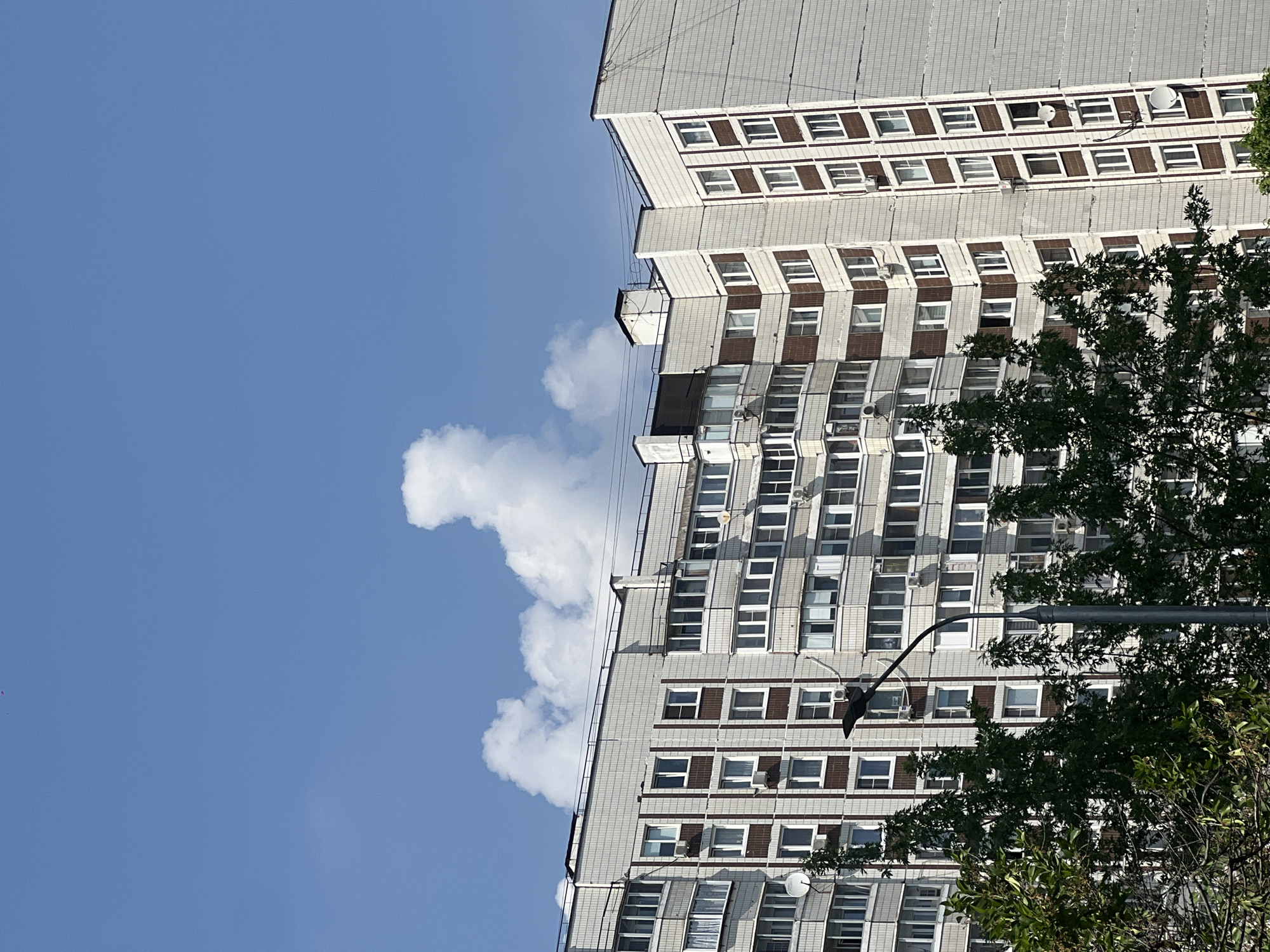 The image depicts a residential building in Moscow, Russia. The building is multi-storied with a grey tiled exterior and rows of windows, some with brown framing. Air conditioning units are visible on some windows. A cloud formation is present beside the building. A tree is located in the foreground. A street light is also visible. The sky is blue and mostly clear.