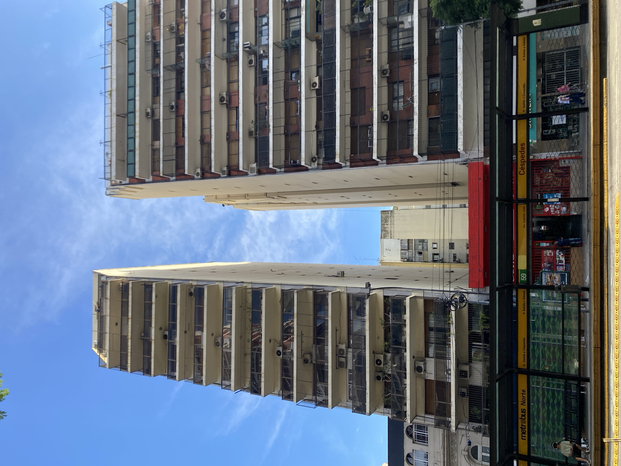The image captures a street scene in Buenos Aires, Argentina, featuring two tall apartment buildings rising high against a clear blue sky. The buildings are characterized by rows of balconies, each adorned with metal railings and air conditioning units.
At the street level, a bus stop labeled "Céspedes" and "metrobus Norte" is visible. Signage indicates directions ("Salida a Céspedes" and "→ Salida a Zabala") and the bus route "59 Av. Cabildo 1200". Two people are sitting on the ground near the bus stop. In the background, a variety of smaller buildings can be seen.