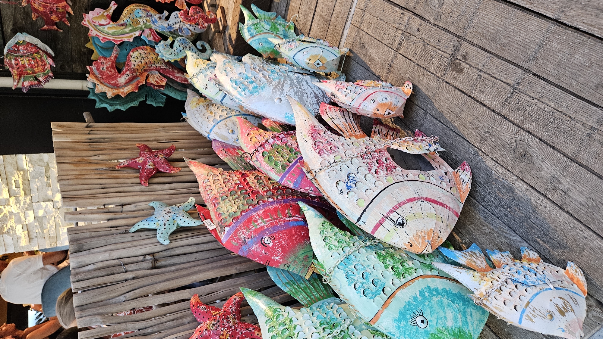 The image displays a collection of colorful, decorative wooden fish and seahorses displayed outdoors on a wooden deck. The fish are varied in size and color, painted with intricate patterns and exaggerated features, giving them a whimsical, folk-art appearance. Some of the fish are suspended by chains, while others rest on the wooden planks. In the background, a weathered wooden fence is visible, with two starfish ornaments hanging from it. Further back, there's a glimpse of a building with a stone facade and people in casual attire, one of whom is seated. The overall impression is that of an outdoor market or shop selling artisanal crafts, possibly in a coastal town. This scene could be located in L'Île-Rousse, France, a town known for its vibrant outdoor markets and coastal charm, where such decorative items are commonly sold as souvenirs.