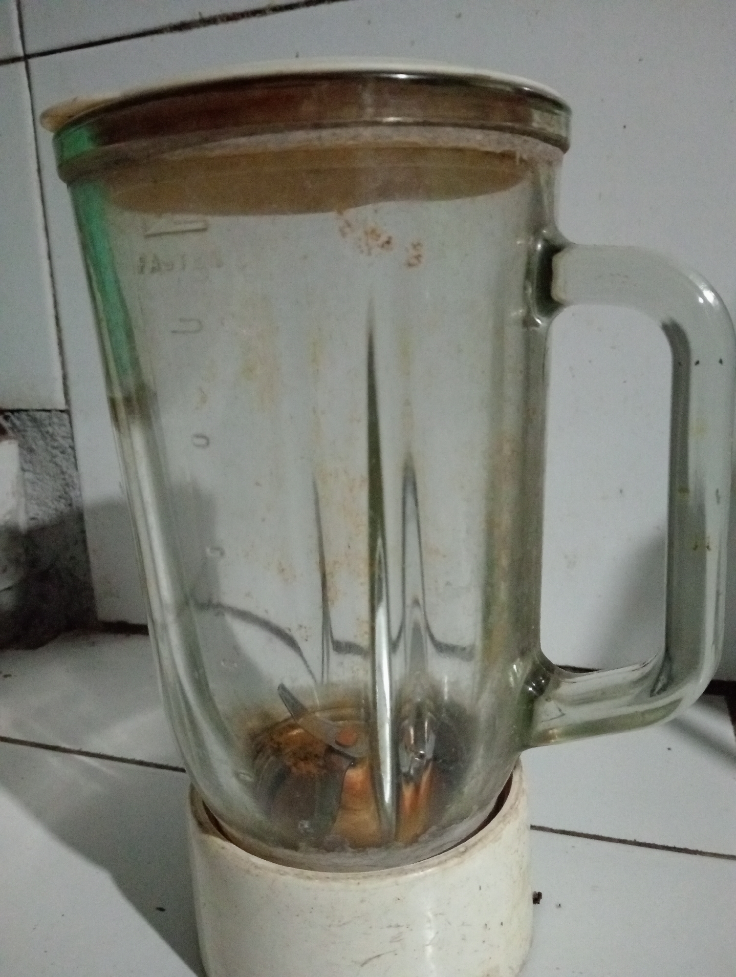 The image shows a close-up of a used glass blender pitcher with a tan lid, resting on a white base. The pitcher is stained and appears to have residue inside and on the exterior. The blender is positioned on a tiled surface, possibly in a kitchen. The lighting is dim, casting shadows. The background consists of white tiles with dark grout lines. The scene is static and focuses solely on the blender. There are no people or actions depicted. The image does not provide any specific information about its location in Trenggalek, Indonesia.