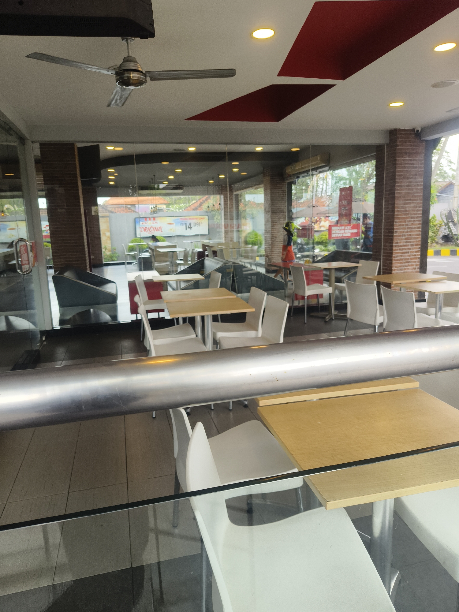 The image shows the interior of a KFC restaurant in Denpasar, Indonesia, viewed from outside through a glass wall and a reflective metal railing. The restaurant features an indoor seating area with multiple tables and chairs. White chairs and light-colored tables are arranged for dining. Beyond the immediate seating area, a red and white sign is visible, likely advertising a promotion. The background reveals more tables and chairs, along with an air conditioning unit mounted on the wall. A ceiling fan is also visible near the entrance. Brick columns and a tiled floor are part of the restaurant's architecture. Outside, trees and a road with a yellow and black striped curb are discernible. One person, wearing a yellow helmet and an orange outfit, is standing inside the restaurant near the sign. The overall scene depicts a typical fast-food dining environment.