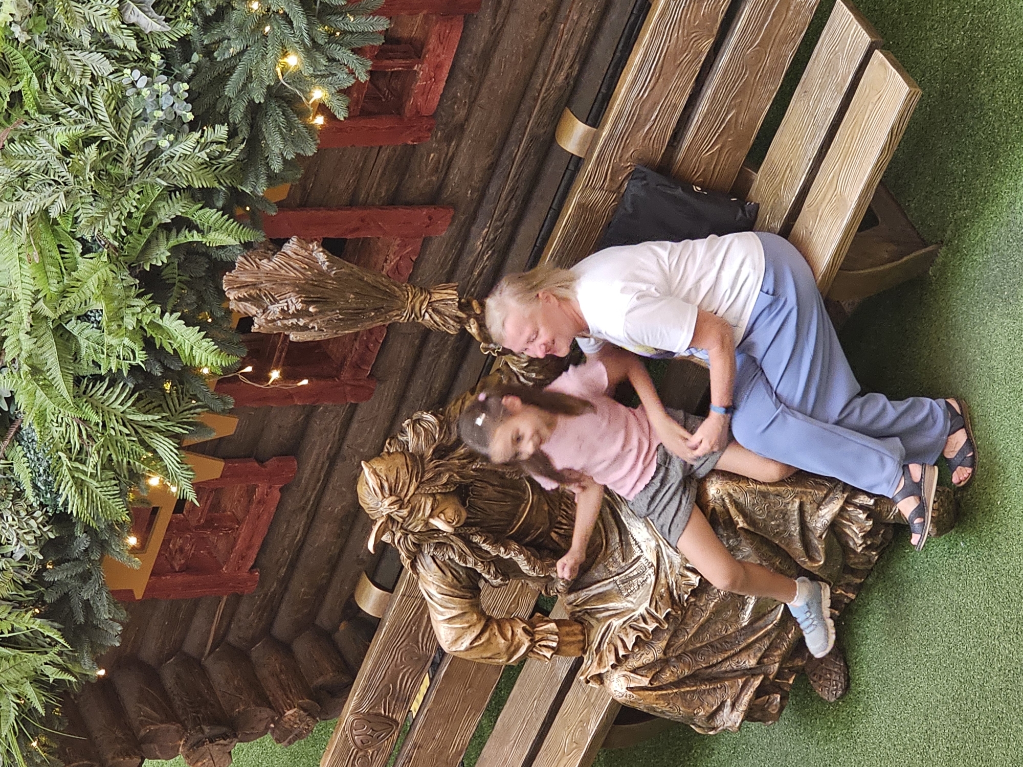 The image shows a bronze statue of Baba Yaga sitting on a wooden bench. A young girl is sitting next to the statue, and an adult woman is sitting next to the girl. They are all on a wooden bench in front of a wooden structure decorated with artificial greenery and lights. The ground is covered in green artificial turf. This scene is located in Moscow, Russia, likely at an entertainment venue or park.