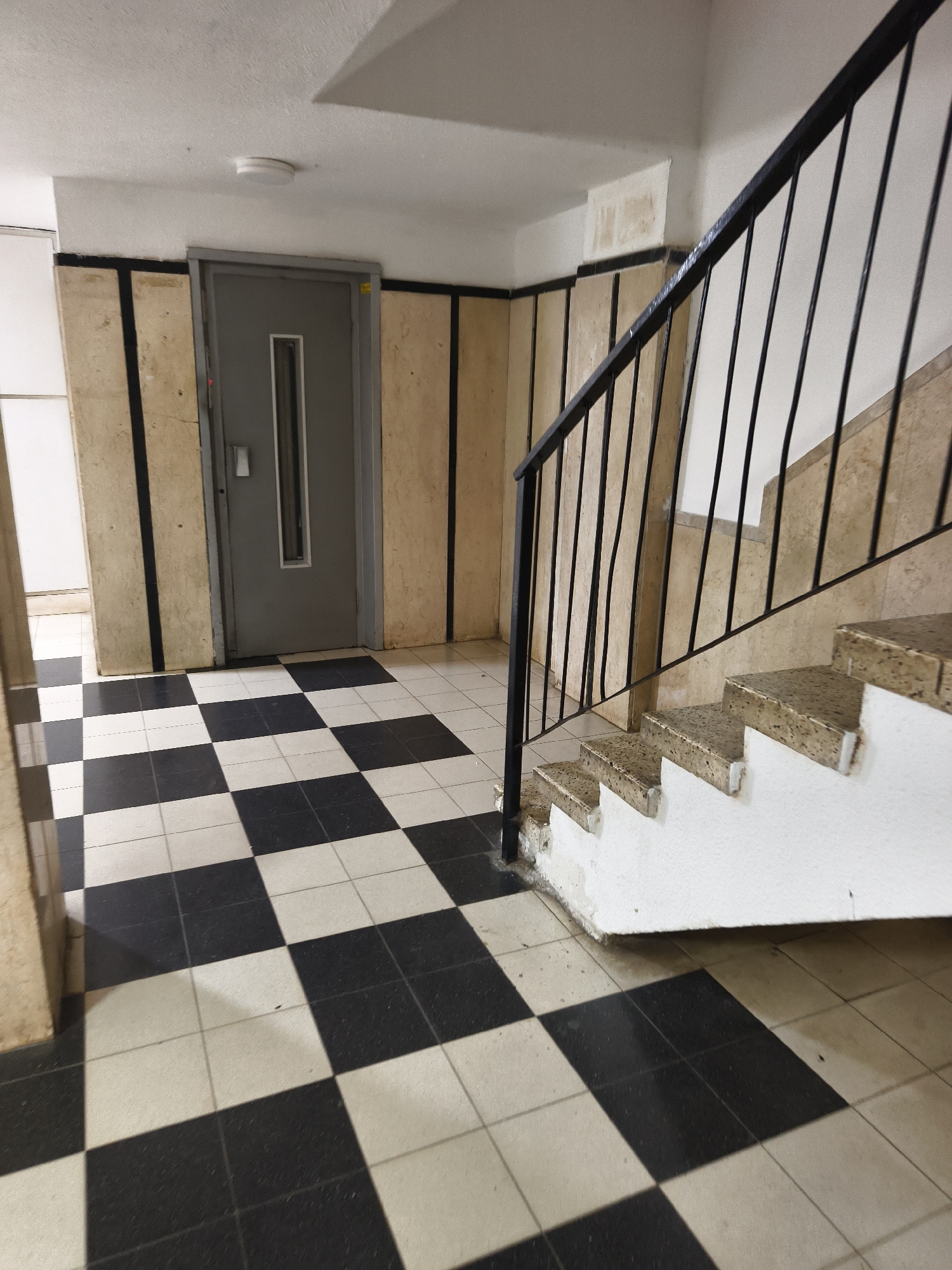 The image depicts the interior of a building, likely an apartment complex, in Petah Tikva, Israel. The scene features a checkered black and white tiled floor, walls paneled in beige with black vertical stripes, and a grey elevator door. To the right, a staircase with a black metal railing ascends. The steps are composed of a speckled beige material with white risers. The overall aesthetic suggests a utilitarian, perhaps dated, design. No people are visible.