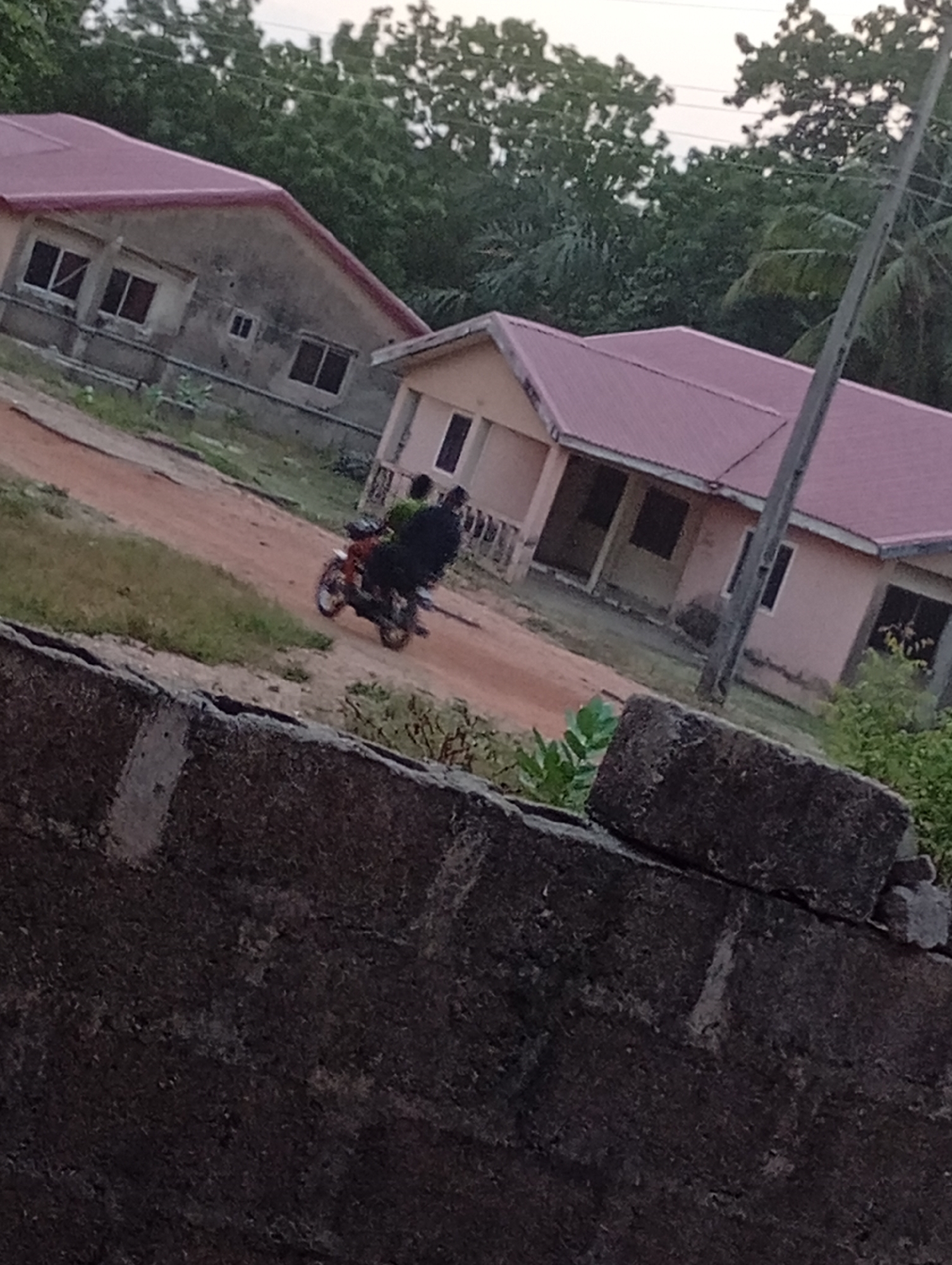 The image depicts a street scene in Unknown, Unknown. Two buildings with red roofs are visible in the background. A motorcycle with two people is driving on a dirt road between the buildings. A rough, dark-colored wall is in the foreground. Trees are in the background behind the buildings. A utility pole is visible on the right.