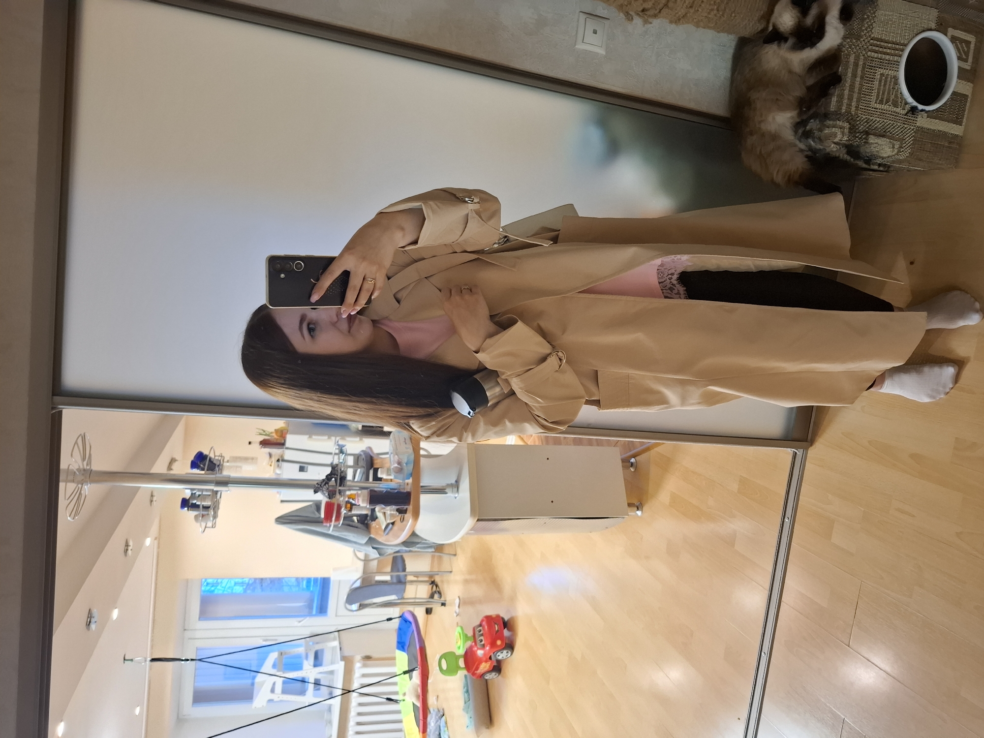 A woman takes a selfie in a mirror, wearing a tan trench coat, pink top and black leggings with socks. The environment reflected includes a salon-style setup with equipment and a child's toys on a wood floor. A cat rests near a cup on a woven mat. The location appears to be an apartment or home in Chaikovsky, Russia.