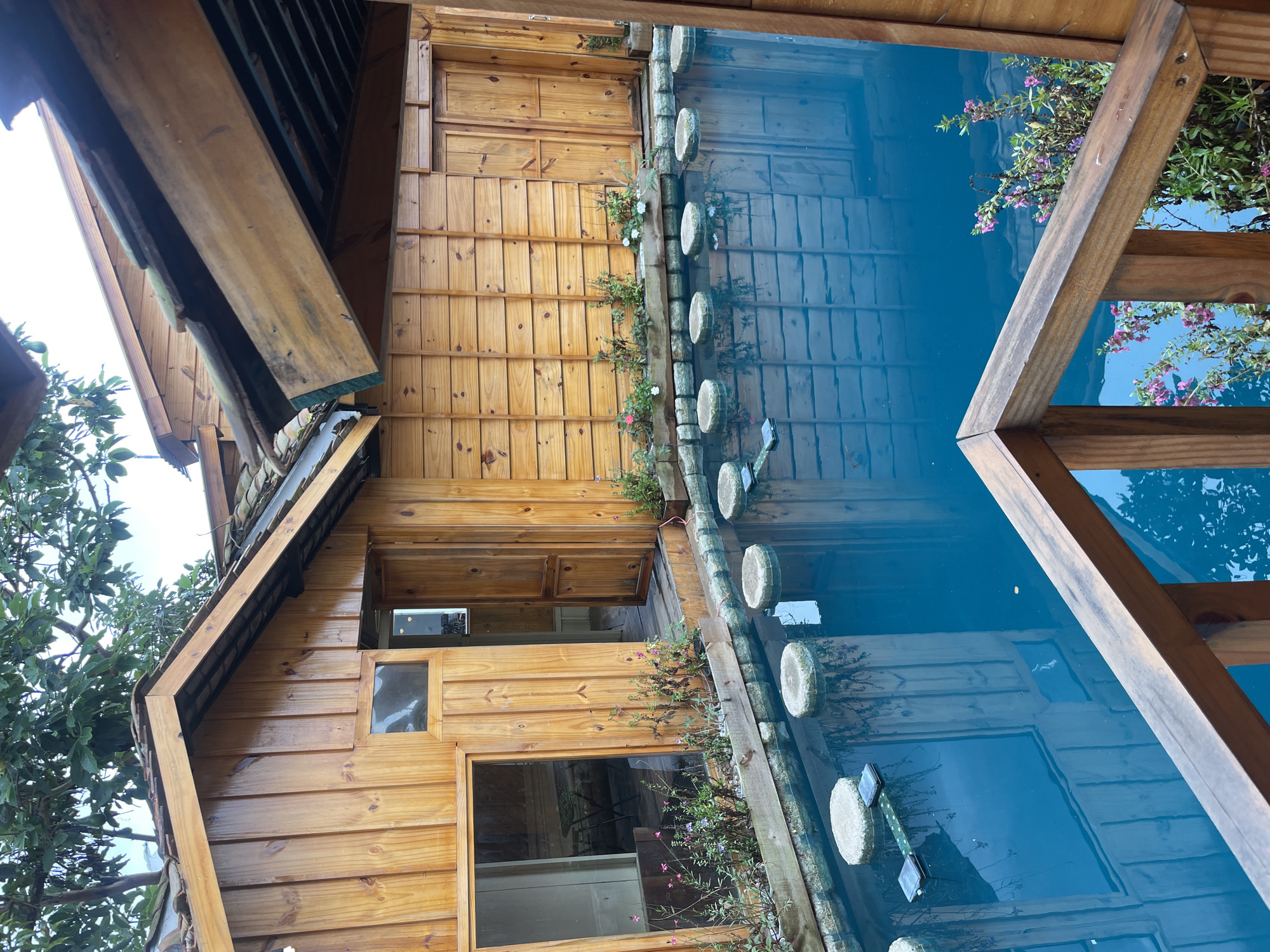 A clear, bright blue pool of water is centrally located in the image, bordered by a wooden deck and stone elements. Stepping stones, round and light gray, are strategically placed across the water, leading towards wooden structures. These structures, built with natural wood, have slatted walls and tiled roofs. One building features a large window with a visible interior containing a table and chairs, suggesting it might be a room or cafe. Another building has closed wooden doors. Small planters with green foliage and delicate pink and white flowers are situated along the edge of the pool. The reflection of the wooden buildings and greenery is visible on the water's surface. Wooden railings frame the foreground, indicating the viewpoint is from a raised platform or balcony. The overall scene suggests a serene, natural setting, possibly a resort or a tranquil residential area in Thôn 3, Vietnam.
