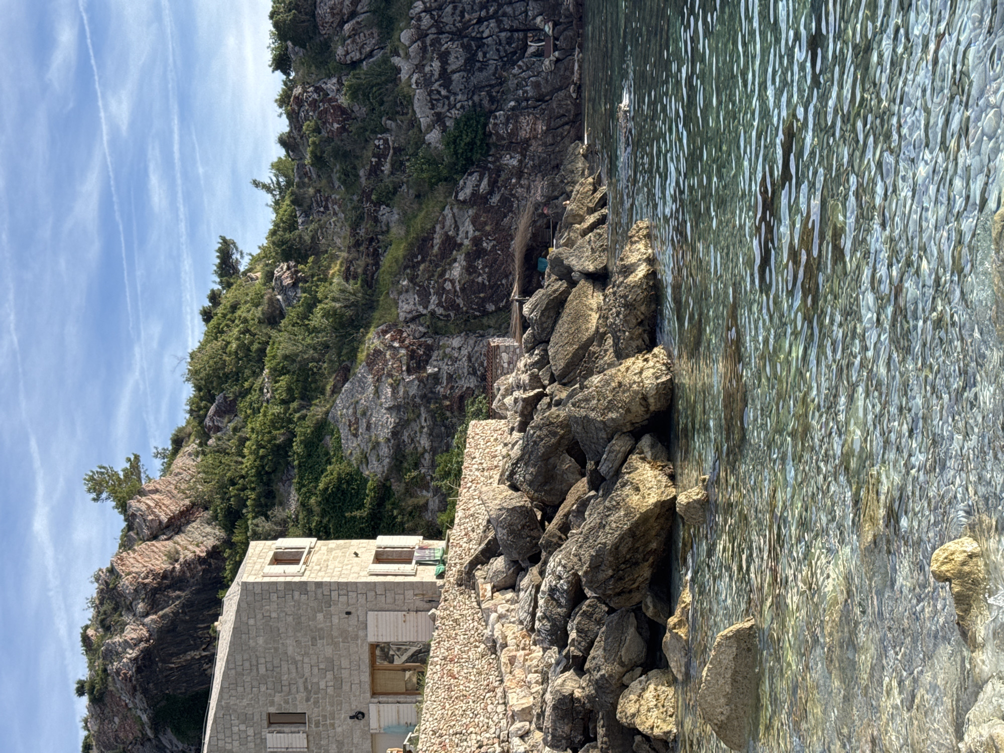 This is a medium-angle, natural light, eye-level shot of a coastal scene in Rijeka Reževići, Montenegro. On the left, a light-colored stone building with white shutters on its windows sits on a rocky slope. A low stone wall, constructed with small, irregularly shaped stones, separates the building from a jumble of large, grey rocks that spill down to the water's edge. The water is clear, revealing a pebbled seabed and reflecting the sunlight. Behind the building, the rocky hillside is covered in dense green vegetation. In the background, a larger, rugged mountain range with exposed pink and grey rock formations rises against a blue sky dotted with thin, white clouds. To the right of the building, further down the coastline, a small, rustic structure with a thatched roof and a fishing net draped over it is nestled amongst the rocks. The scene depicts a quiet, natural coastal environment with a touch of human habitation.