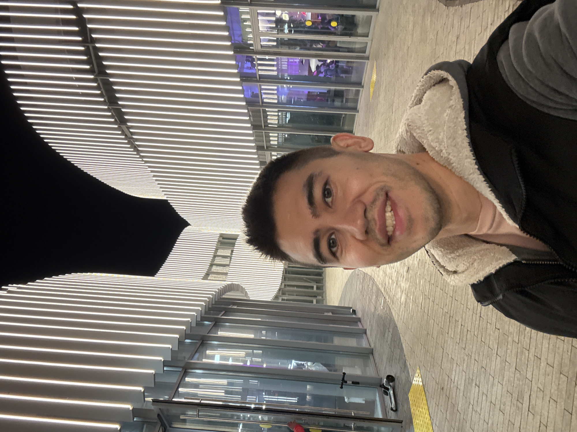 A smiling man takes a selfie. He wears a black hoodie with a white sherpa-lined hood over a pink t-shirt. Behind him is the Kazan Family Center, a building known for its unique cauldron shape and decorative lighting. The exterior of the building is visible through glass windows, indicating the man is likely standing outdoors. The background also shows part of the textured pavement and architectural lighting of the plaza. The image appears to be taken at night or during twilight, given the dominance of artificial light.