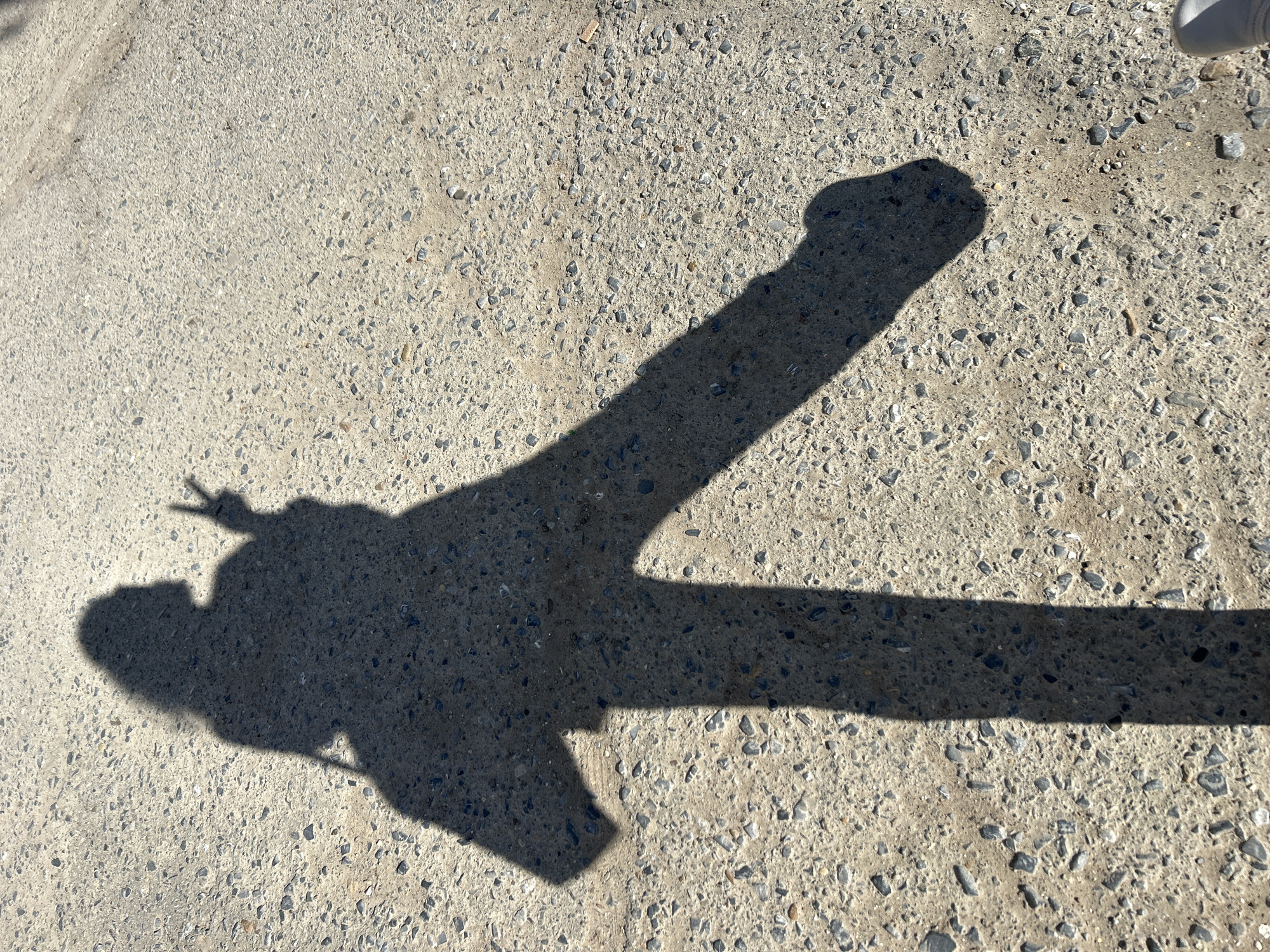 The image shows a shadow on a paved surface. The shadow is of a person holding their arm out and fingers in a peace sign. The location is outdoors, likely on a sidewalk or road in Chaikovsky, Russia. The paved surface is light gray and appears to be made of concrete with small pebbles or aggregate visible. The lighting indicates it is daytime and sunny. There is also a part of a shoe in the top right corner.