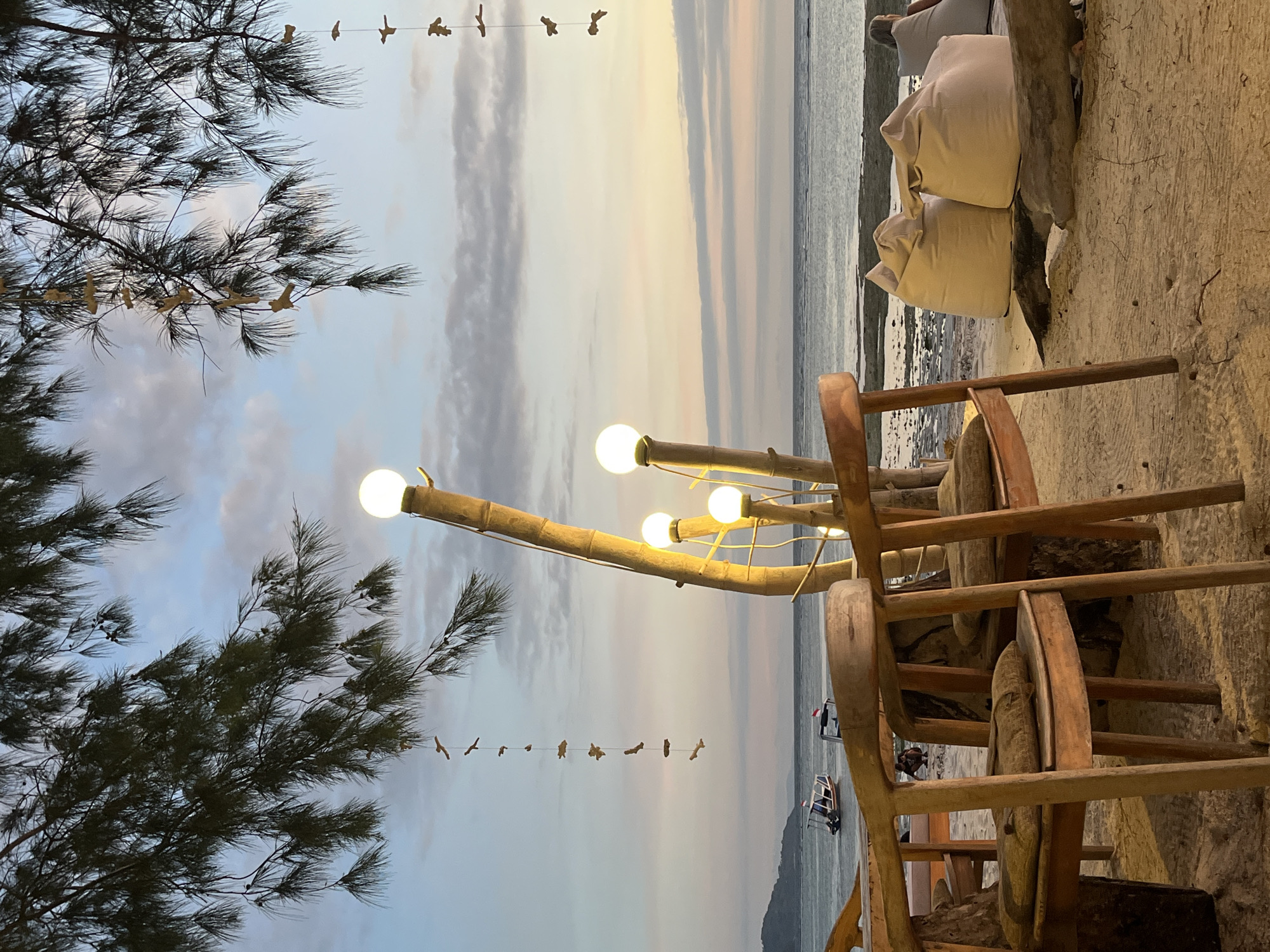 The image captures a beachside scene in Unknown, Indonesia, presenting a blend of natural and human-made elements. The composition is dominated by three wooden chairs in the foreground, hinting at a relaxed, informal seating arrangement. The beach's sandy terrain is visible at the base of the chairs, with a raised concrete edge separating the seating area from the shoreline.
To the left, a tree with fine, dark foliage frames the view, adding a natural boundary to the scene. A string of small, bird-shaped ornaments hangs delicately between the tree and a set of bamboo light fixtures. These fixtures, equipped with spherical bulbs, provide ambient lighting, suggesting a late afternoon or early evening setting.
In the background, the sea meets the horizon under a sky painted with hues of blue, white, and a hint of yellow, indicating either sunrise or sunset. The presence of beanbag-like cushions stacked behind the chairs further reinforces the relaxed, beachside atmosphere. The distant view reveals a small island or landmass and boats in the ocean, adding depth to the scene.