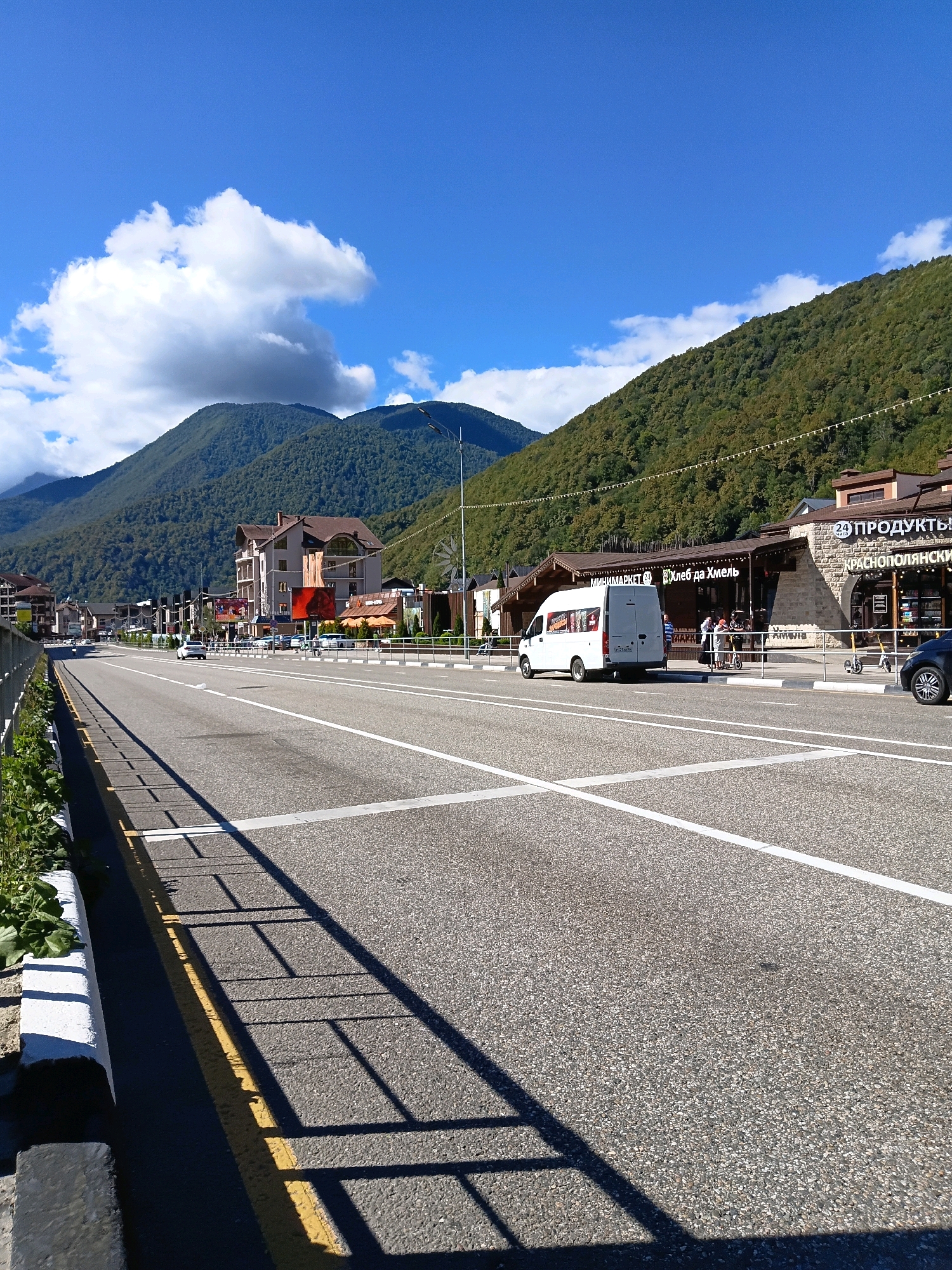 This is a wide-angle, eye-level, outdoor, daytime photograph taken on a sunny day in Estosadok, Russia. The scene depicts a straight, multi-lane asphalt road lined with white and yellow markings. On the right side of the road, there are several low-rise buildings with brown roofs and stone facades, appearing to be shops or businesses, with awnings and signage. Some of these signs have Russian text, including "ПРОДУКТЫ" (Products) and "МИНИМАРКЕТ" (Minimarket). A white van is parked on the side of the road, and several people are standing near it and in front of the shops. Two yellow electric scooters are also visible. A dark-colored car is parked on the far right. The road is bordered by a white curb and a metal fence on the left. Lush green trees cover the steep mountains that rise behind the buildings. The sky is a clear, bright blue with a few white, fluffy clouds. The overall impression is that of a resort town or a commercial area situated in a mountainous landscape.