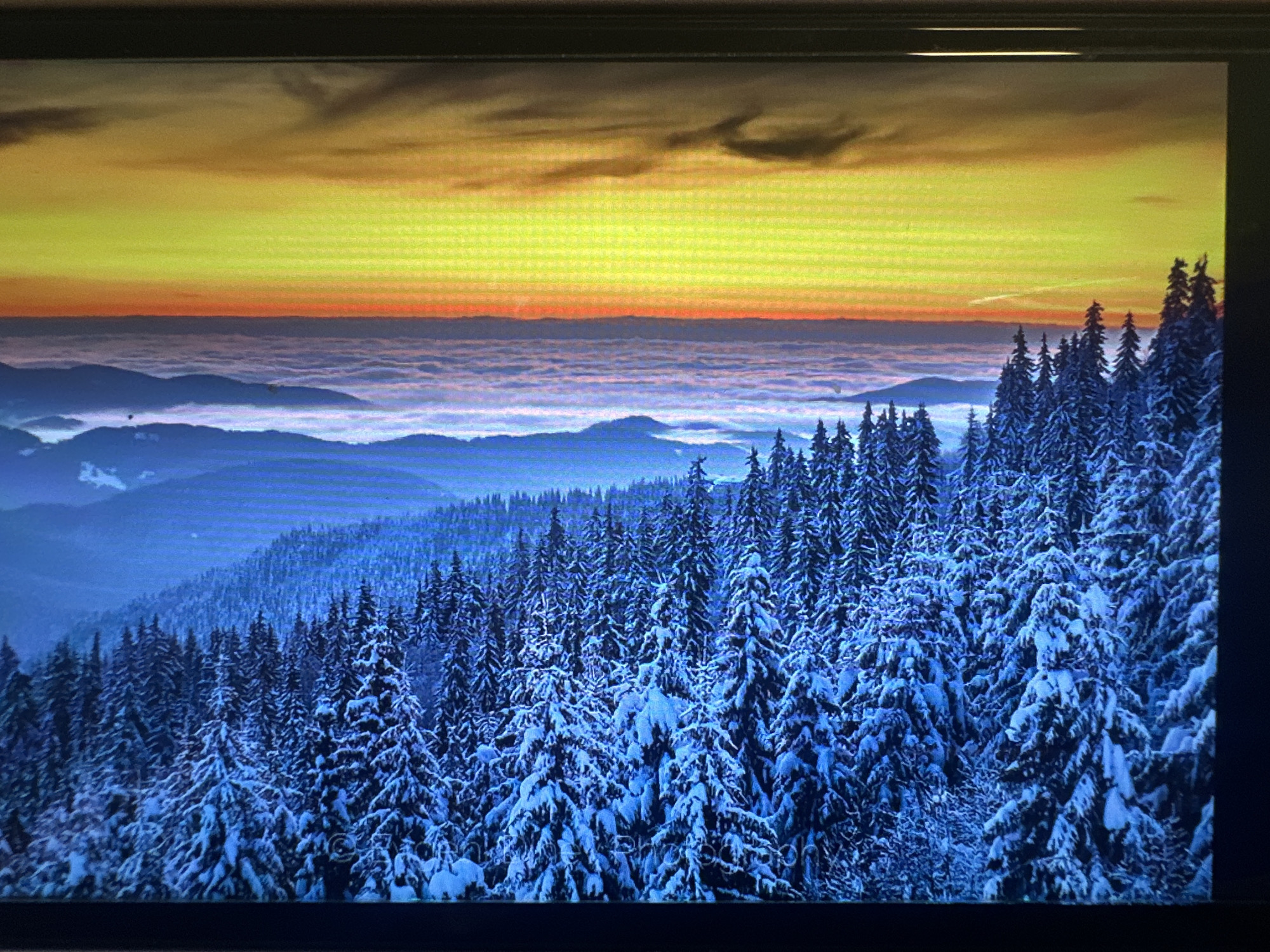 The image displays a digitally rendered scene of a winter forest with snow-covered evergreen trees on the left, transitioning to a vibrant sunset or sunrise with clouds and a bright yellow and orange sky on the right. The perspective is from above, looking down and across the landscape. There are no people, animals, or specific landmarks visible that would indicate a location in the city of Mainburg, Germany. The scene appears to be a high-resolution digital wallpaper or artwork.