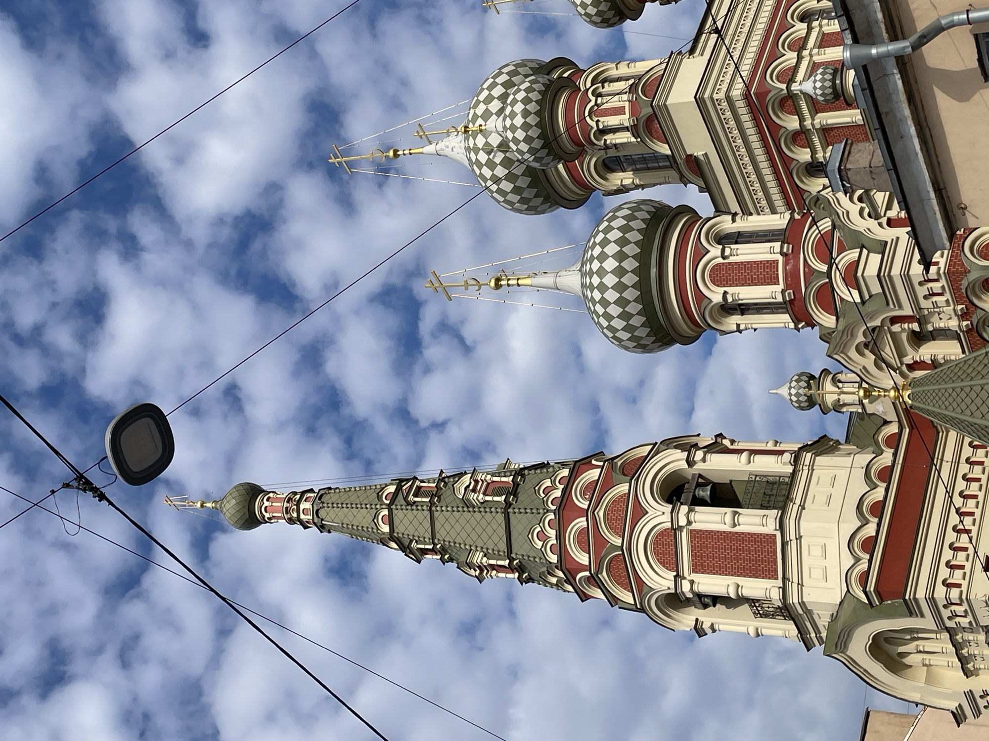 The original image, presented rotated 90 degrees clockwise, captures a detailed view of a Russian Orthodox church against a bright, cloudy blue sky in Saint Petersburg, Russia.
The primary subjects are the church's elaborate architectural elements. Several onion-shaped domes are prominent, characterized by a distinctive green and white checkered pattern, each topped with a golden Orthodox cross. A tall, slender spire rises towards the sky, adorned with intricate geometric patterns in muted green tones and also culminating in a cross. The church building itself showcases ornate detailing, with creamy white walls, rich red brickwork accents, and decorative arched windows and cornices. An open section of a bell tower, revealing bells within, is also visible. The architectural style is highly decorative and richly colored.
In the foreground, dark utility lines crisscross the frame, partially obscuring the view. A modern, dark grey street lamp or overhead light fixture hangs from these wires. On the far right, a small section of a building's roof with a gutter is visible, suggesting the photo was taken from an urban environment, looking upwards.
The bright sunlight and scattered white clouds against the blue sky indicate that the photograph was taken during daytime. There are no people visible, and no discernible text is present in the image.