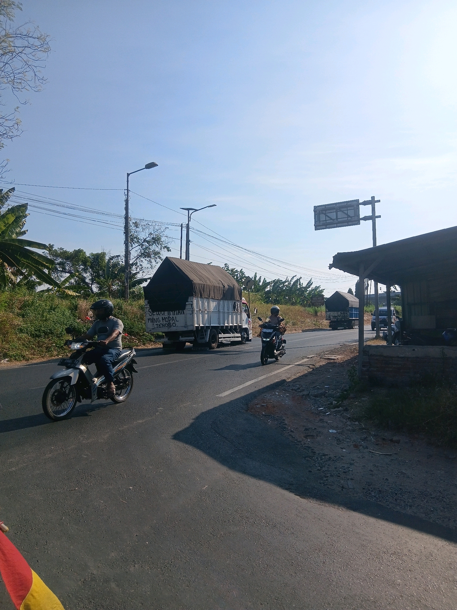 This is a wide-angle, eye-level, outdoor photograph taken in Gedeg, Indonesia, on a clear day with a bright blue sky. The scene depicts a road with vehicles and surrounding vegetation.
In the foreground, a motorcycle is being ridden on the right side of the road. The rider, wearing a gray t-shirt, blue jeans, and a black helmet, is partially visible. The motorcycle is white and silver with black wheels. A shadow of the motorcycle and rider falls across the asphalt road.
Further down the road, a white truck with a dark tarp covering its cargo is also traveling. Text is visible on the side of the truck, reading "SEWA TIRIM" and "MING METAL TANOGO."
To the right of the road, a rudimentary open-air structure with a dark roof made of corrugated material is present. Brick pillars support the roof. Beyond the structure, more vegetation is visible, including banana trees and various bushes.
In the background, more trucks can be seen on the road, and power lines crisscross the sky, supported by utility poles. A road sign with a white rectangular surface is mounted on a pole. The overall impression is a sunny day on a road in a somewhat rural or semi-urban area of Indonesia.
