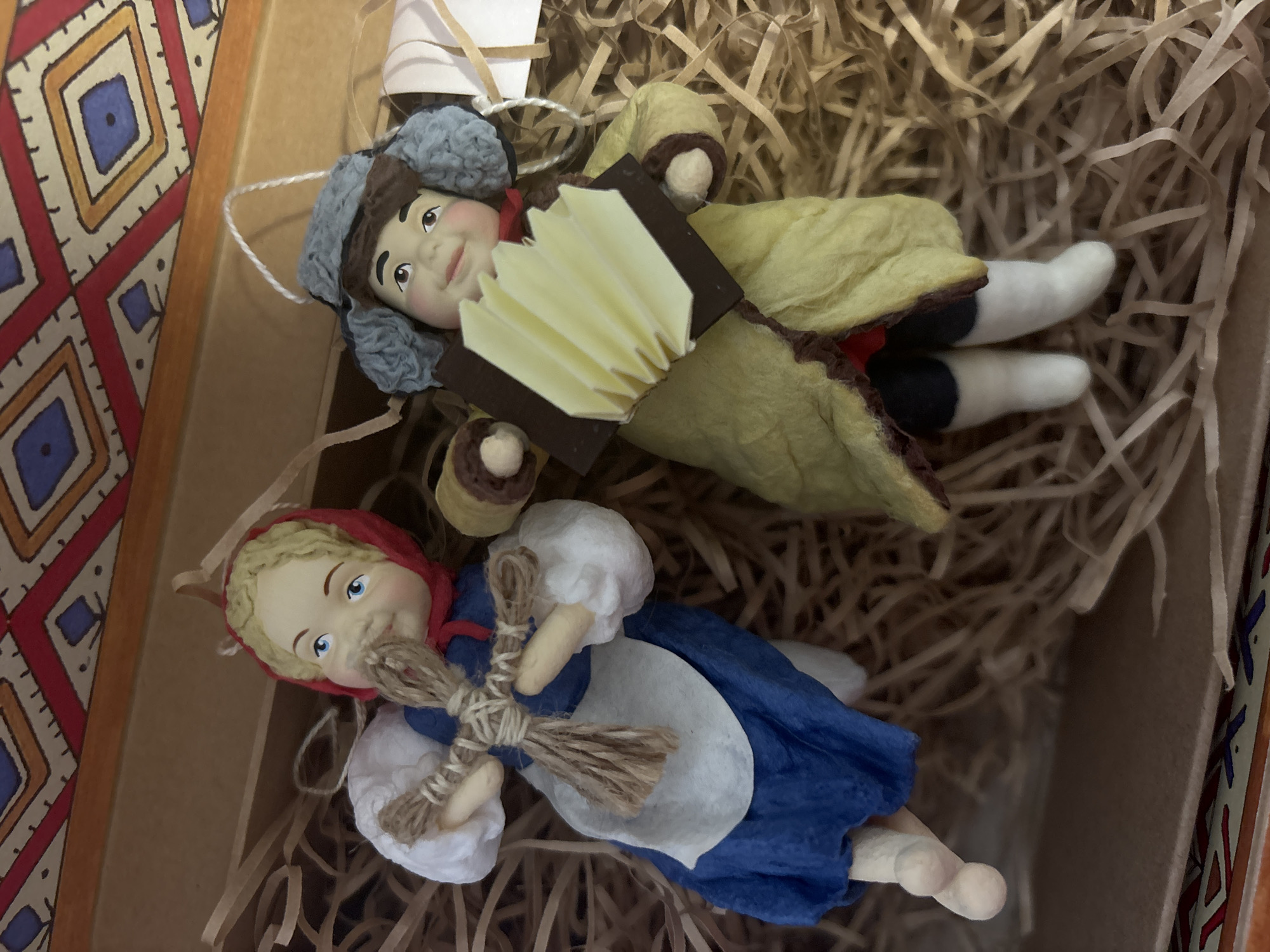 Two handmade dolls lie in a box filled with shredded paper. The doll on top is male, wearing a blue fur hat and a long, yellow coat with brown trim. He holds a small, rectangular accordion with off-white keys. The doll below is female, wearing a red headscarf, a blue dress, and a white apron. She holds a bundle of twigs tied with string. The box appears to be part of a larger set or collection, with patterned borders visible along the edges. The setting is possibly related to the city of Chaikovsky, Russia, suggesting the dolls may be traditional Russian folk figures or characters.