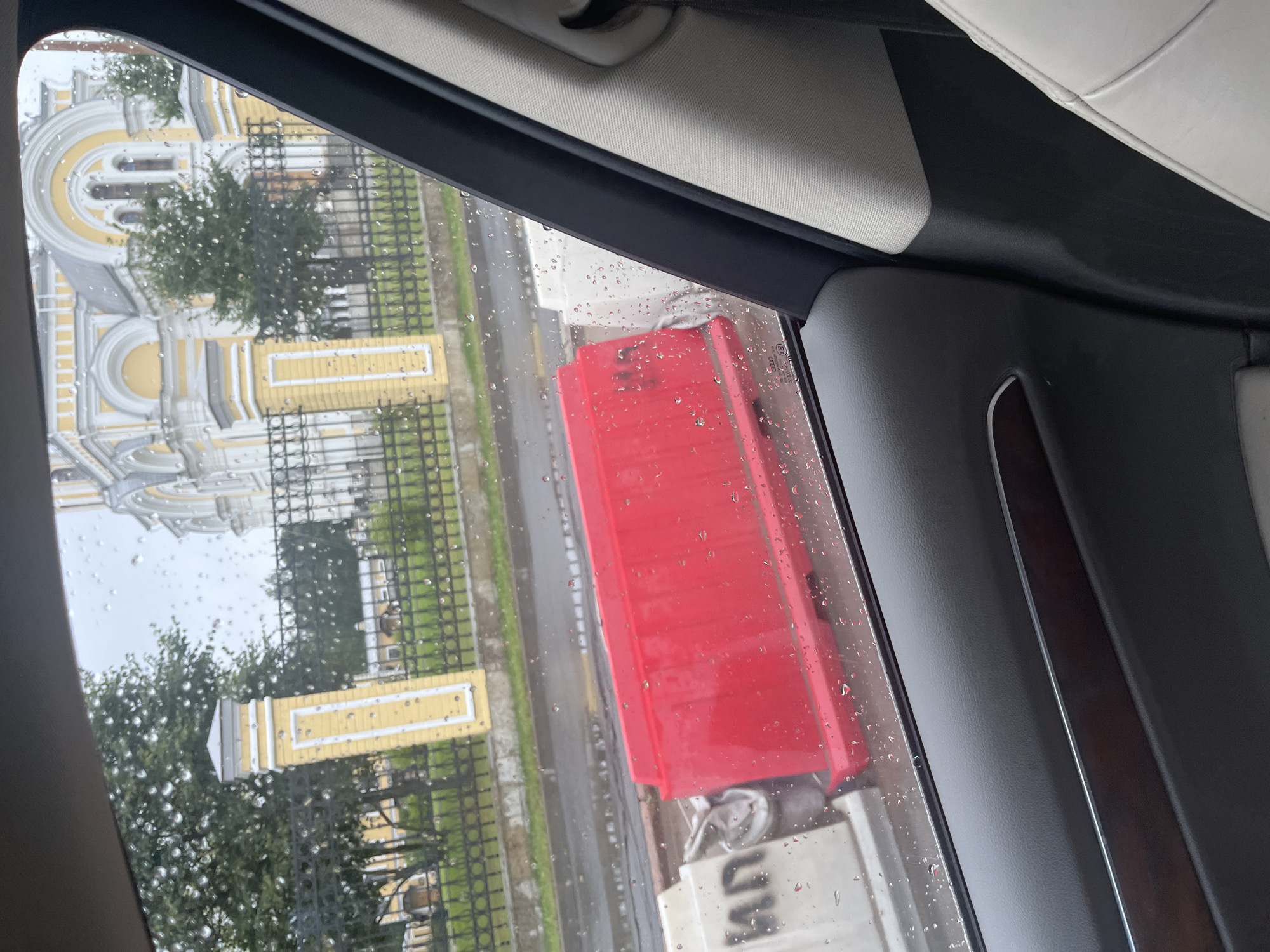 The image, taken from inside a vehicle, captures a scene through a rain-streaked window. Outside, a yellow building with ornate white architectural details, possibly a church or public building, stands behind a black metal fence. The weather appears overcast, with visible raindrops on the windowpane. In the foreground, red and white construction barriers are visible on the side of a wet road. The interior of the car shows a dark grey door panel with a wood-grain accent and a light-colored seat. The context suggests a temporary obstruction or roadwork in an urban setting. While the specific location within Saint Petersburg is not explicitly identifiable from the visual information alone, the architectural style of the building is consistent with that found in the city.