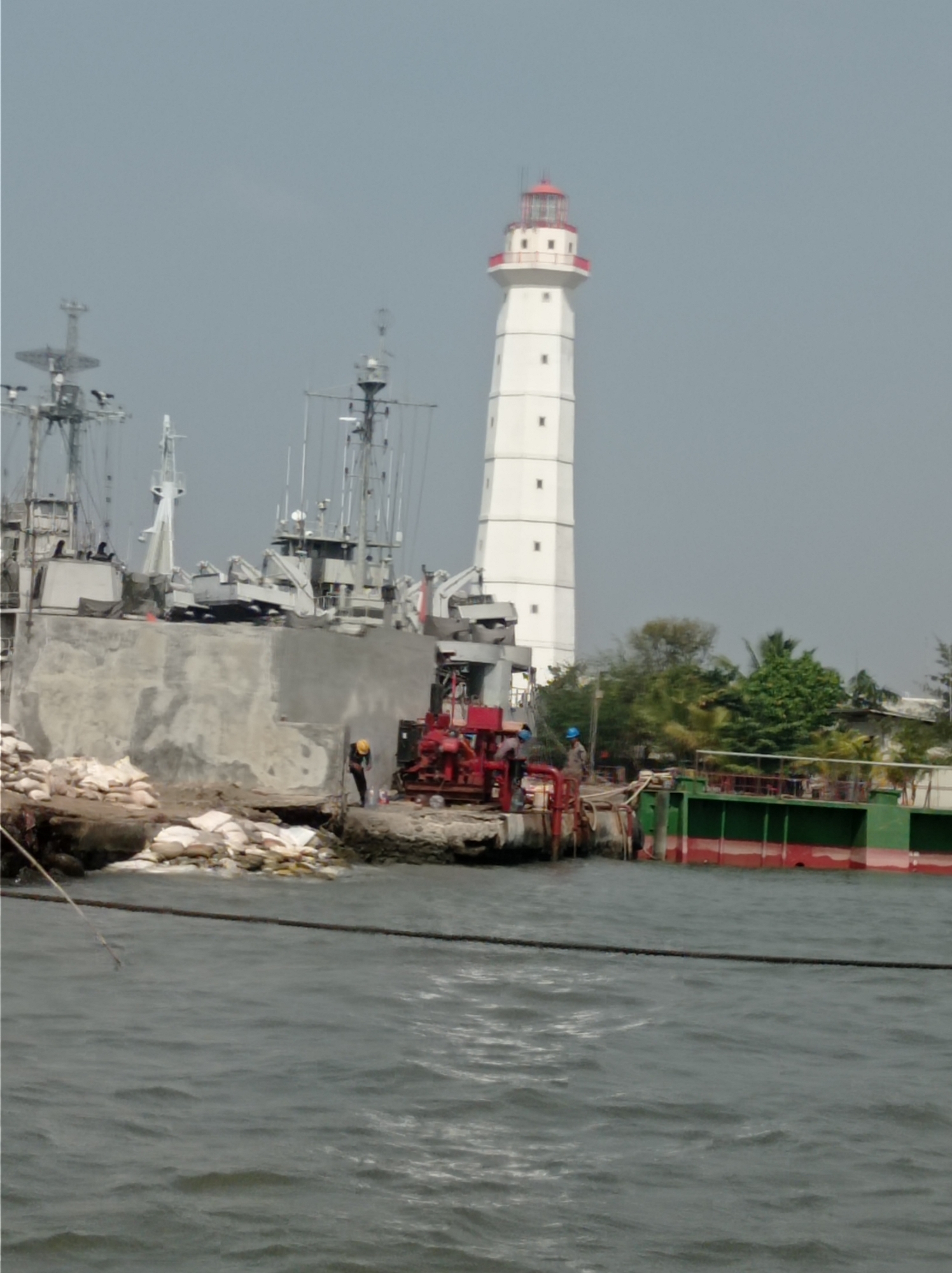 The image depicts a harbor scene in Tegal, Indonesia, dominated by a tall, white lighthouse with red accents. In the foreground, the dark water of the harbor is visible, with a thick rope stretched across it. To the left, several ships, likely naval vessels due to their stacked superstructures and masts, are moored. A large, concrete structure, possibly a seawall or dock extension, forms the midground. On this structure, a few individuals in work attire, one wearing a yellow hard hat, are present, along with a red construction vehicle and sandbags. To the right, a green and red barge is docked. Lush green trees can be seen behind the harbor structures. The overall impression is one of an active port with ongoing construction or maintenance activities.