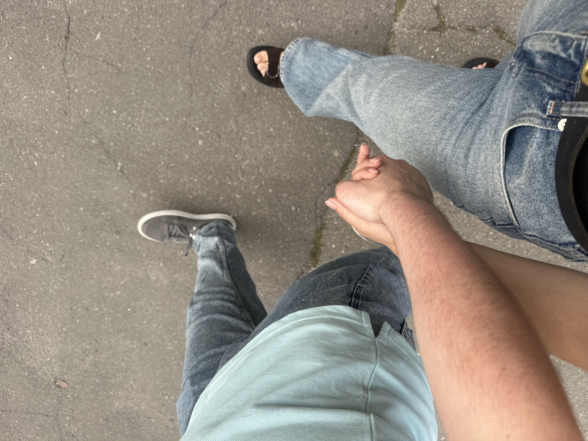 An elevated, ground-level, over-the-shoulder shot captures two individuals walking hand-in-hand on an asphalt surface, consistent with a street or walkway in Ternopil, Ukraine. The person on the left wears a light blue polo shirt and blue jeans, their right arm extended to hold the hand of the person on the right. The person on the right is dressed in light blue jeans and dark sandals, their feet pointing away from the camera. The asphalt shows signs of wear, with small pebbles and subtle cracks visible. The background is nondescript, focusing on the interaction between the two people.