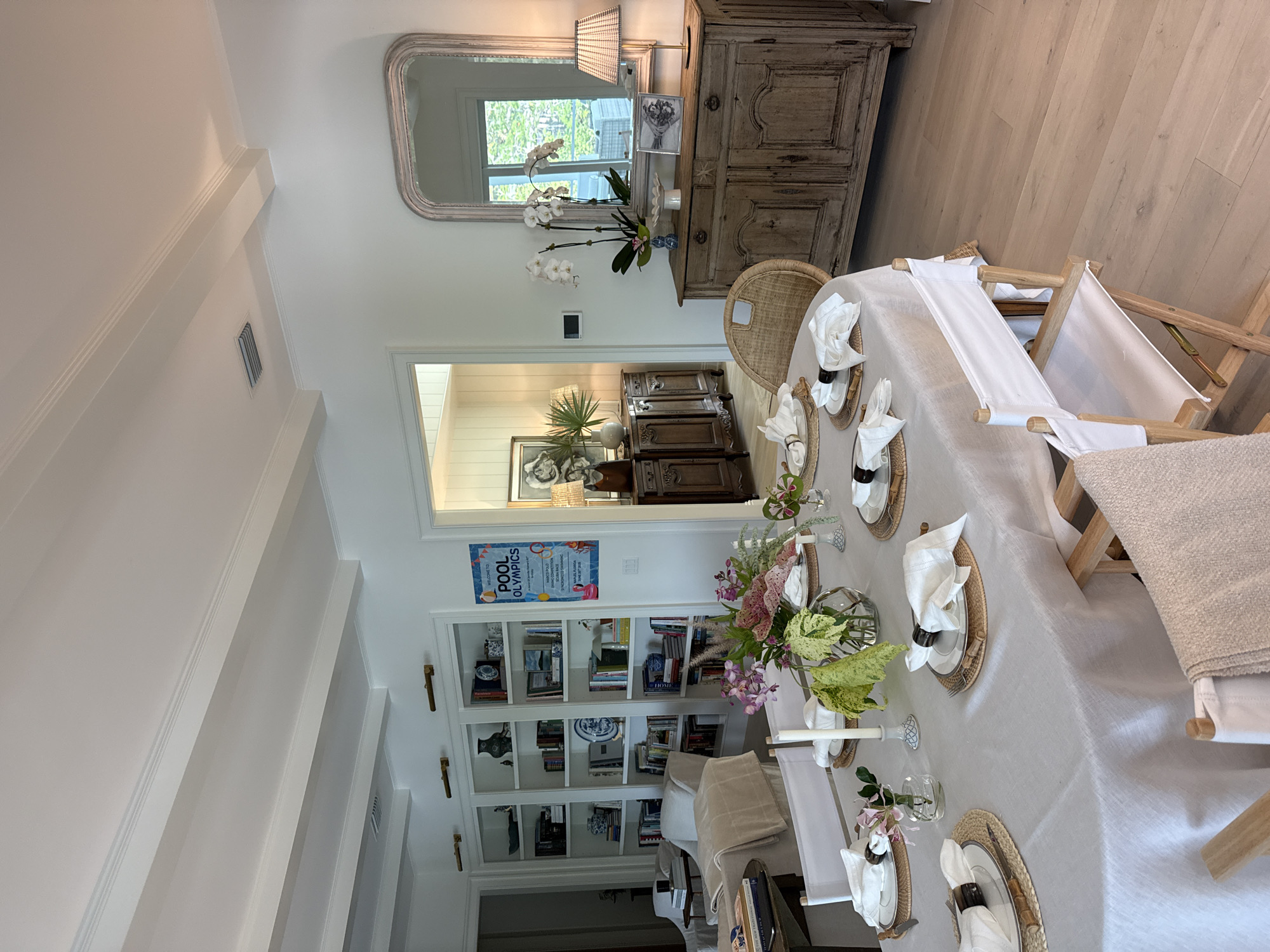 The image depicts an interior scene of a dining room, likely in East Naples, Florida, given the style of the architecture and decor. A large, oval dining table is centrally positioned, covered with a light-colored linen tablecloth. The table is set for a meal with placemats, plates, white napkins with dark napkin rings, and cutlery. A floral arrangement in a glass vase and several candles adorn the table. White director's chairs with light-colored canvas seats are placed around the table.
In the background, a white wall features a large built-in bookshelf filled with books and decorative items. To the right of the bookshelf, a framed "POOL OLYMPICS" poster is displayed. Further to the right, a distressed wooden sideboard stands against the wall, topped with a large ornate mirror. On the sideboard, there are decorative objects including a lamp with a pleated shade, a vase with orchids, a white bowl, and a framed photograph. Through an open doorway to the left, another room with light-paneled walls and a wooden cabinet is visible. The room has a light-colored wood floor and a coffered ceiling with white beams. No people are present in the image.