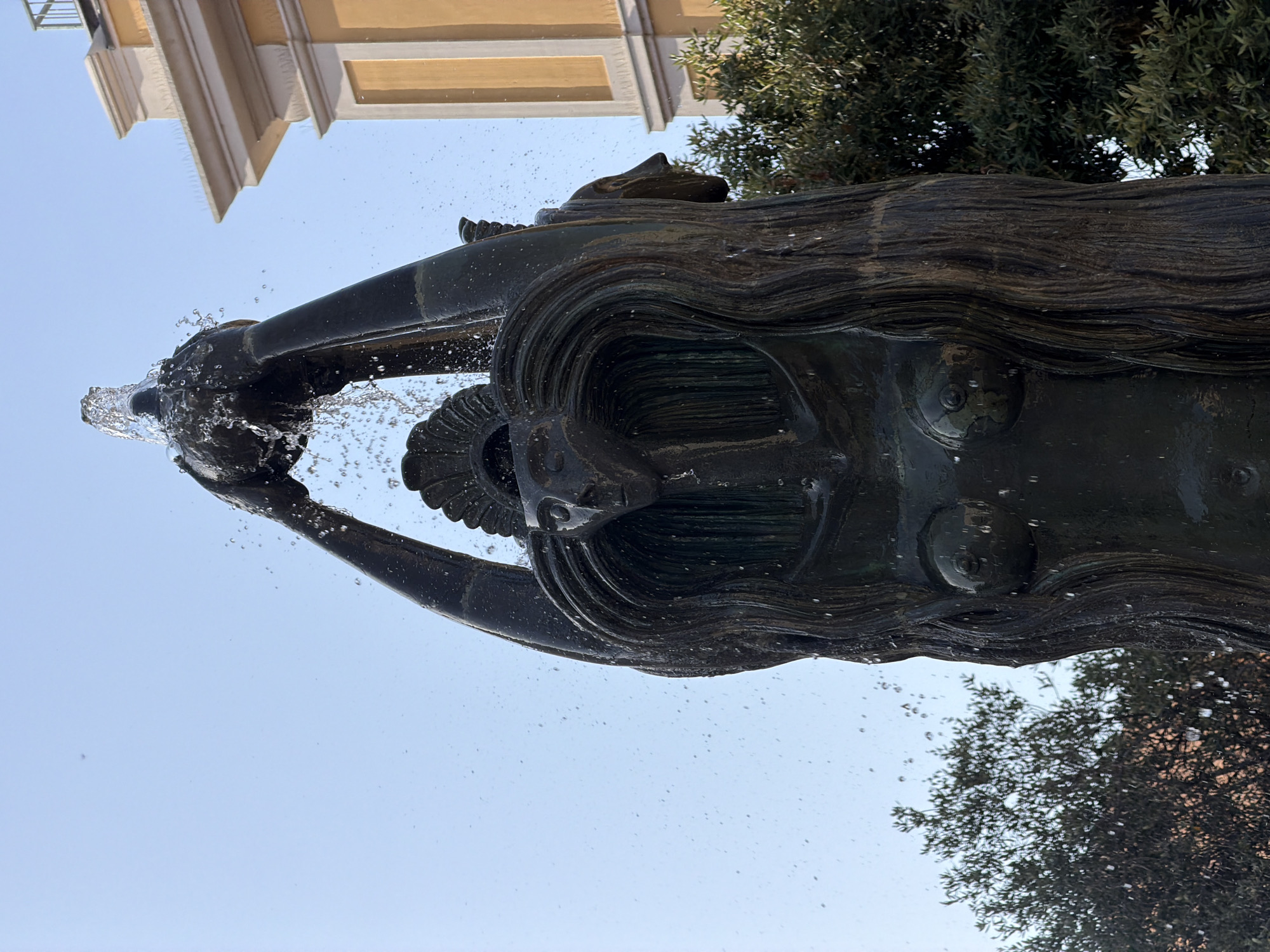 The image shows a bronze fountain sculpture in the city of Olbia, Italy. The sculpture depicts a female figure with long, flowing hair and a headdress. Water is spraying from a vessel held above her head. The sculpture is set against a clear blue sky with a building and trees visible in the background. The building appears to be yellow with white architectural details.