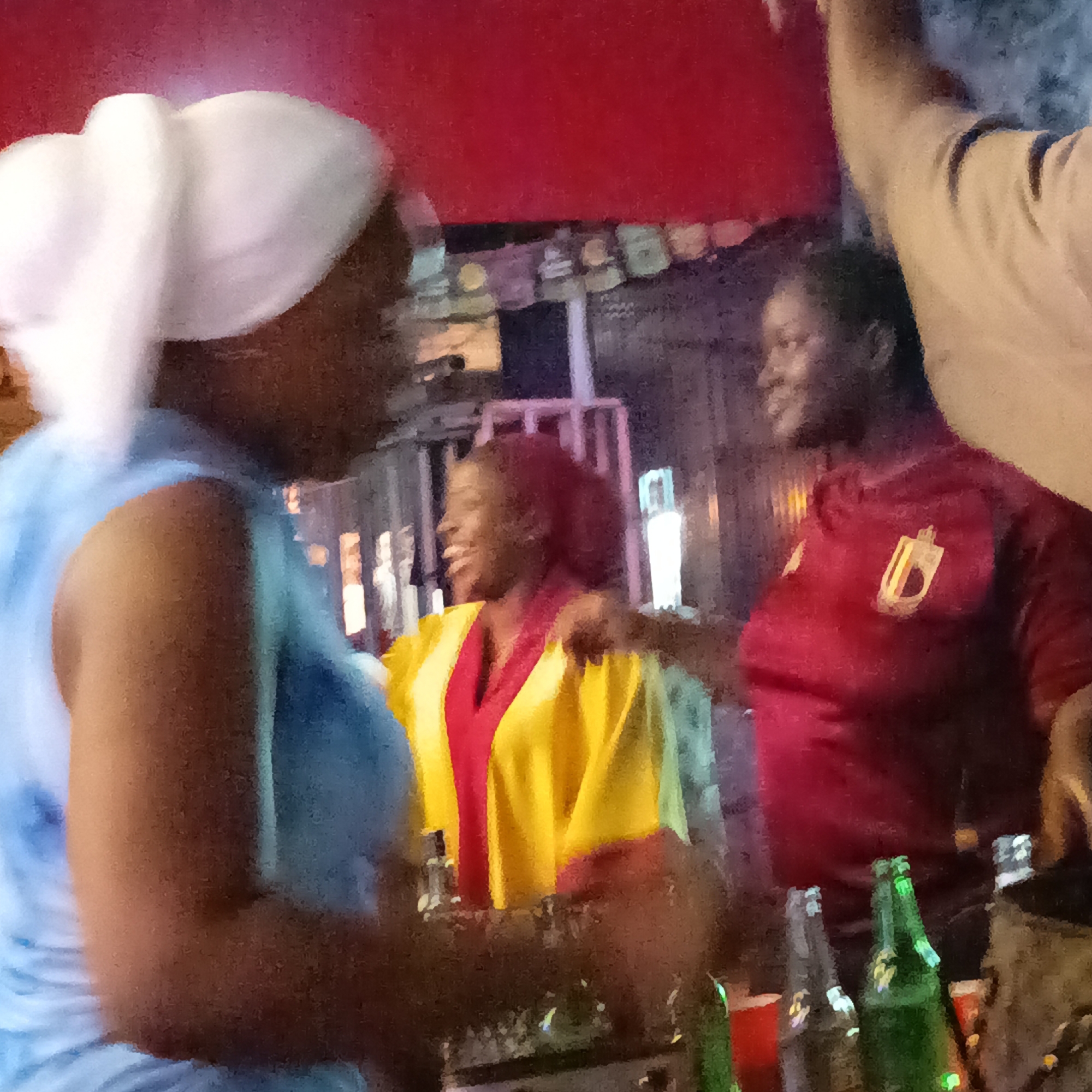 The image shows three Black women socializing at an outdoor bar or event in Akwanga, Nigeria. The woman on the left is wearing a light blue sleeveless garment and a white head wrap. The woman in the center is wearing a yellow robe or jacket with a red accent. The woman on the right is wearing a red jersey with a gold logo. In the foreground are assorted bottles of beer and plastic cups on the bar. In the background, there is an indiscernible structure decorated with colorful flags, with additional architectural features suggestive of a built-up urban environment. Overhead, a section of a red awning is visible.