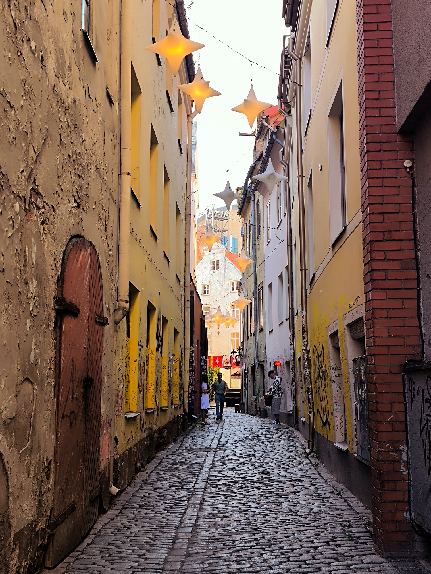 The image depicts a narrow cobblestone alleyway in Riga, Latvia, lined with buildings on either side. The architecture suggests an old European city, with some buildings featuring weathered facades and others painted in bright colors. Star-shaped decorative lights hang across the alley, indicating a festive or seasonal display.
On the left, a building with a textured, peeling facade has a large, rustic wooden door. The building on the right is a mix of painted surfaces and exposed red brick. Graffiti is visible on the walls of several buildings, adding a layer of urban character.
Two individuals are walking down the alley. One, a male in a green shirt and jeans, is walking towards the viewer. Another, a female in a white dress, is also walking away from the viewer. Further down the alley, another person, possibly male, is wearing a striped shirt and shorts and appears to be interacting with something on the ground or a low surface.
The cobblestone street is uneven, typical of historic European city centers. The overall impression is of a charming, slightly aged, and atmospheric urban passage. The presence of the star lights suggests it might be a scene during a holiday season or a special event.