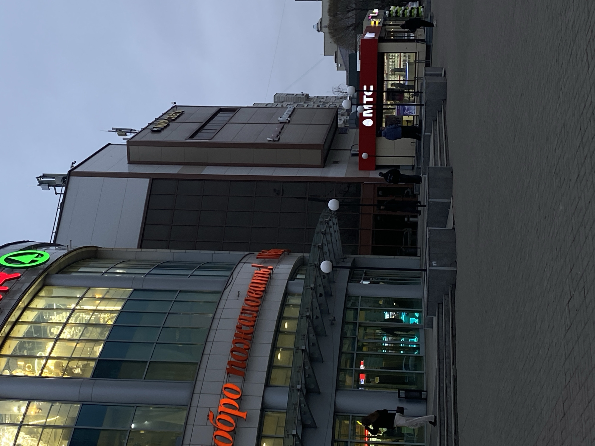 The image shows a street scene in Kazan, Russia, with a view of several buildings. The most prominent building on the left has a curved glass facade and displays the Russian words "Добро пожаловать" in orange lettering. On the building's roof, there is a green circular logo. Adjacent to this building is a taller, rectangular building with darker, tinted windows. The name "Suvar Plaza" is visible on the top of this building. To the right, a red vertical sign with the letters "MTC" is present, indicating a mobile telecommunications store. Several people are visible on the sidewalk, some walking and some standing. The ground is paved with grey bricks. The sky is overcast, suggesting it is either dusk or dawn.
