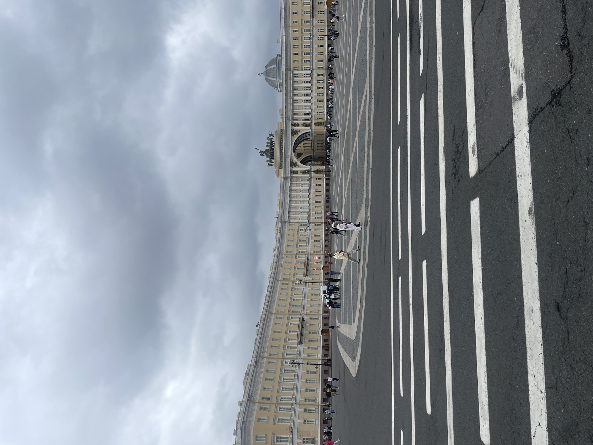 The image shows a wide, paved square with pedestrian crossings in the foreground. In the background, a large, pale yellow, Neoclassical building complex dominates the scene, featuring a prominent central archway topped with a chariot sculpture. The building has numerous windows and a curved facade. The sky is overcast with gray clouds. Several people are scattered across the square, walking and standing. A white van is parked near the building. The location is Palace Square in Saint Petersburg, Russia.