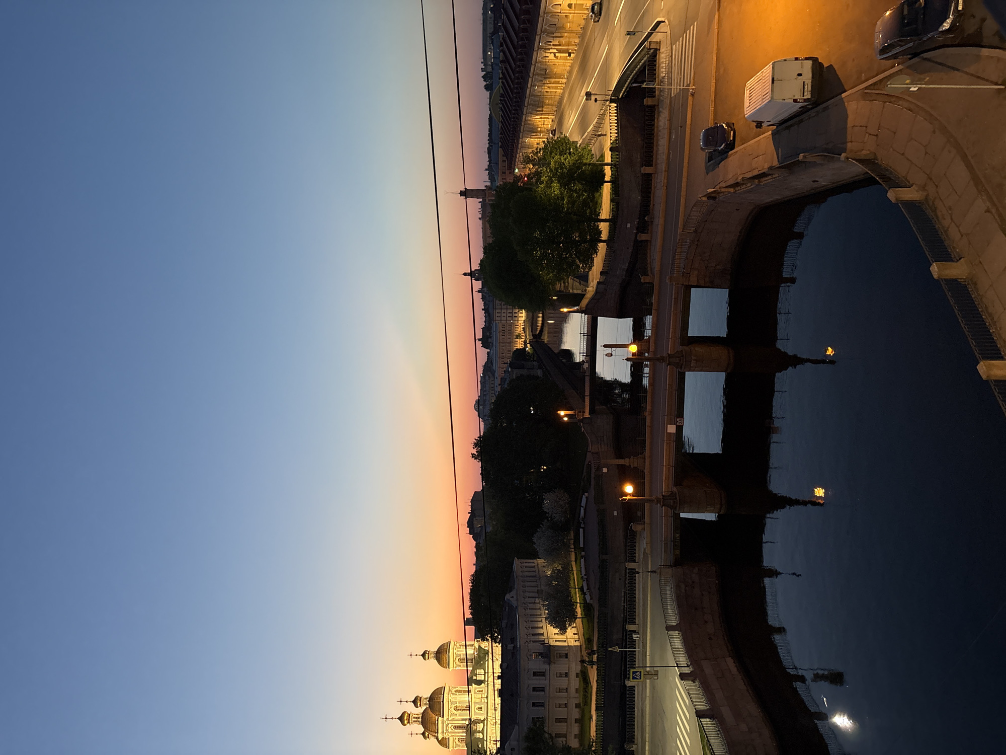 A night view in Saint Petersburg, Russia shows a canal, likely the Moika, under a stone bridge. The sky transitions from blue to orange near the horizon, where buildings, including a church with golden domes, are illuminated. Trees and streetlights are visible along the canal banks. Cars are parked and driving on the bridge. The canal water reflects the lights. Power lines stretch across the sky.