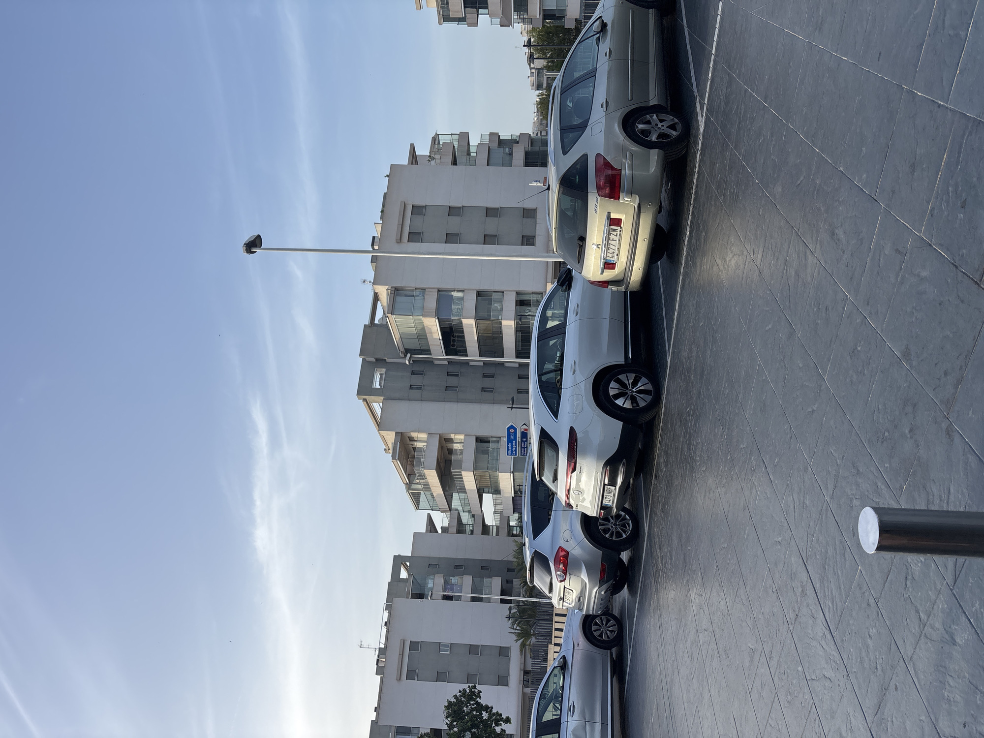 The image depicts a parking lot in Urbanización Villa Martín, Spain, with several cars parked in designated spaces. The cars are lined up facing away from the viewer, and include a silver station wagon, a silver SUV, a white crossover SUV, and a white electric vehicle. In the background, there are modern apartment buildings with multiple balconies. A streetlamp stands tall on the right side of the image. The parking lot surface is paved with large, dark grey tiles. The sky is clear and blue with a few wispy clouds, suggesting it is daytime and the weather is fair. The overall scene is quiet and orderly.