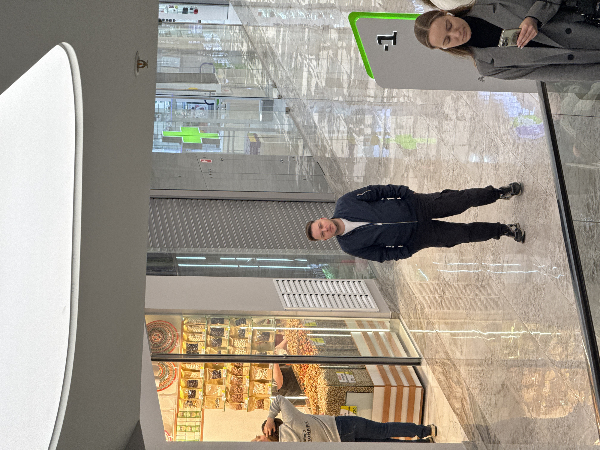 The image captures an indoor scene in a shopping mall in Moscow, Russia. The focus is on a glass display case filled with dried fruits and nuts, likely from a store specializing in such products. Two individuals are visible near the display case: one person is taking a photo of the display, while another person is observing or shopping nearby.
Further in the background, beyond the glass display, a man is standing. Beside him a woman is also present, engrossed in her phone.
The shopping mall has a modern interior with gray-tiled floors. The presence of signage and other retail stores in the background suggests a busy commercial environment.