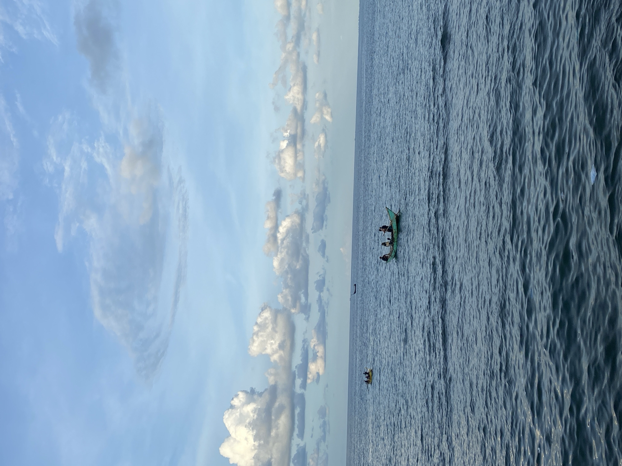 The image depicts a coastal scene, likely in Lhokseumawe, Indonesia, featuring a cloudy sky over the sea. Two boats, one with visible occupants, are on the water. The sea is a dark blue-gray and appears moderately choppy. The image is rotated 90 degrees clockwise.