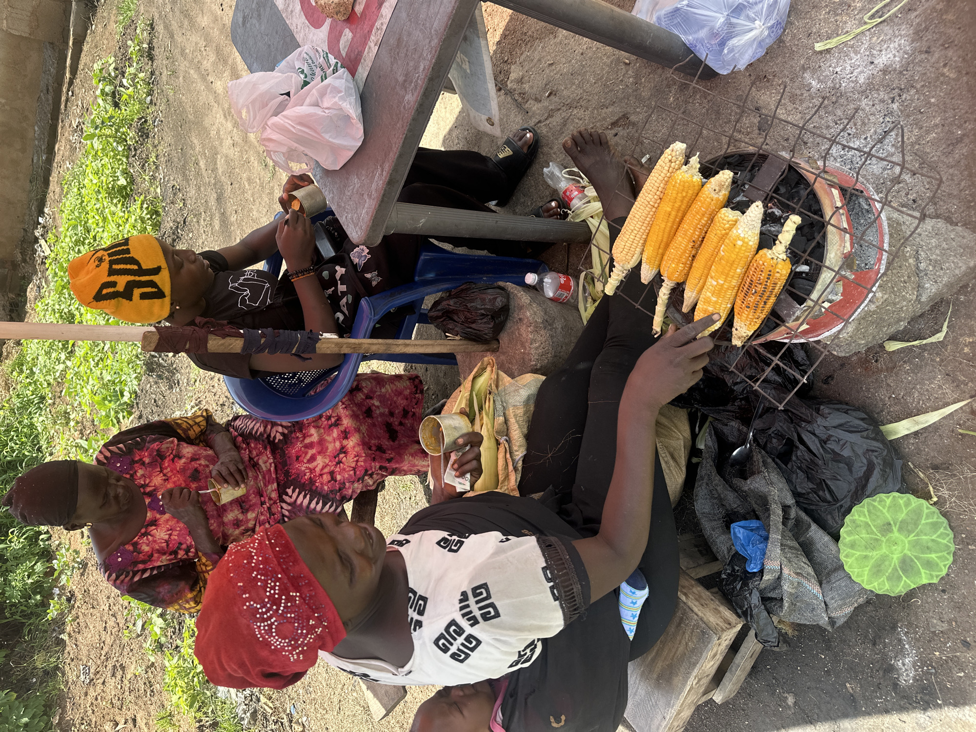 In Gura Top, Nigeria, a woman wearing a red headscarf is preparing corn on a grill. She is seated next to a baby carrier holding a baby. Next to her, another woman in a colorful dress is eating from a cup. A child in an orange beanie sits at a table, also eating from a cup. The scene appears to be an outdoor food stall, with corn cobs arranged on a metal grill over coals. Various plastic bags, bottles, and a green decorative item are scattered around the ground. The environment suggests a sunny day with vegetation in the background.
