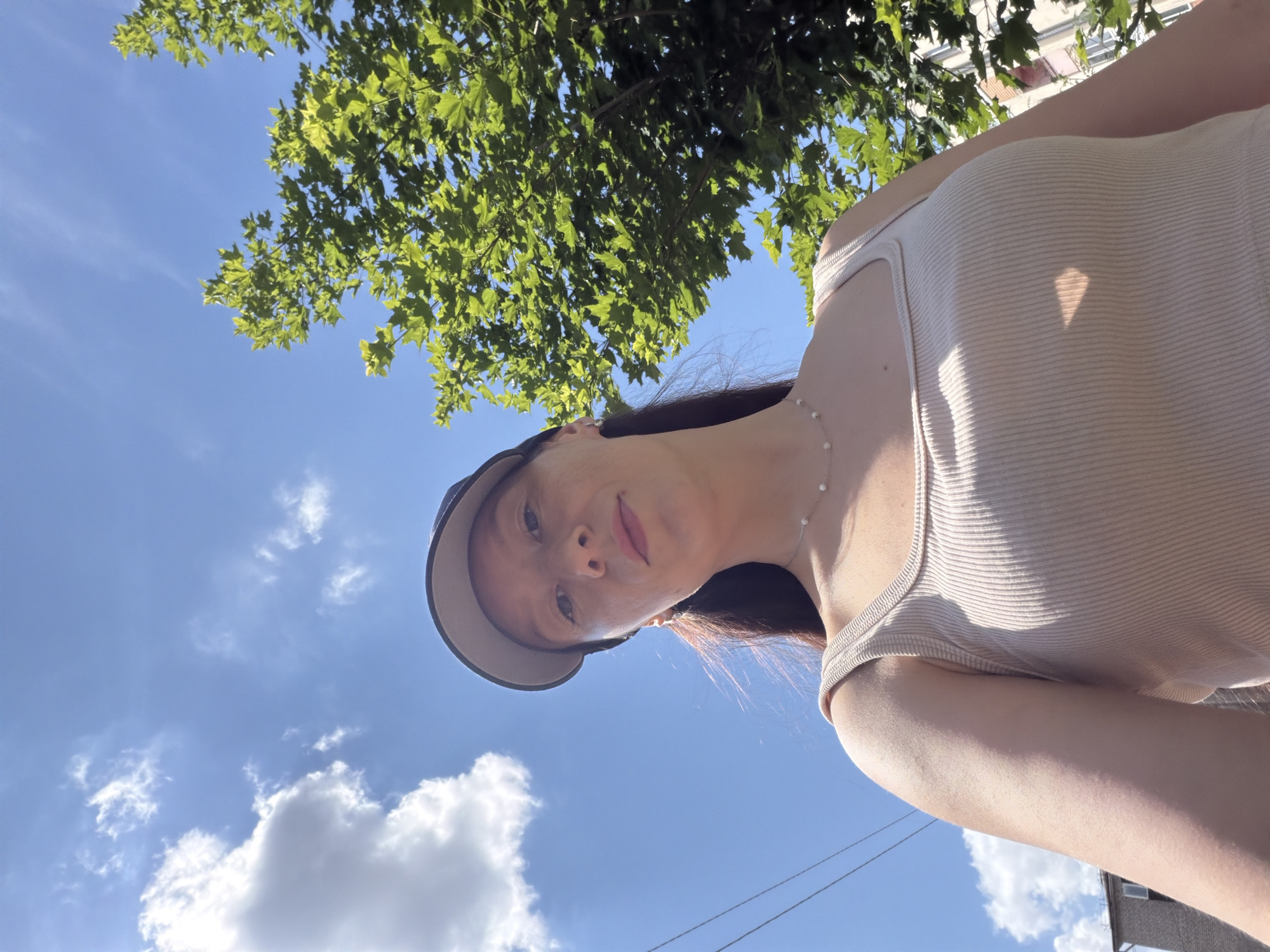 A low-angle, medium shot captures a woman from the chest up, facing the camera and looking slightly upwards. She is wearing a beige ribbed tank top and a dark baseball cap. A delicate silver necklace with small white beads adorns her neck. Her hair, dark brown, is pulled back into a ponytail. Behind her, the bright blue sky is dotted with fluffy white clouds. To the right, a lush green tree with vibrant leaves is visible, its branches reaching towards the sky. The background also shows the upper portion of a brick building and power lines, suggesting an urban setting. The lighting indicates a sunny day.