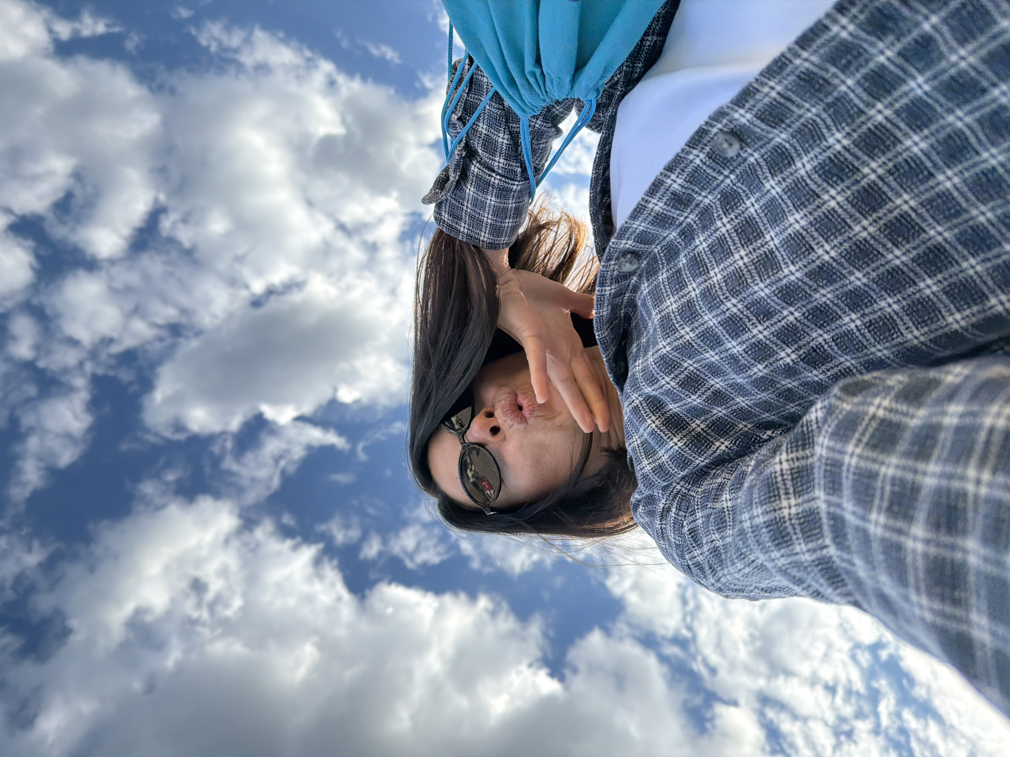 A woman with dark hair is looking downward at the camera. She is wearing sunglasses and a blue and white plaid shirt. She is holding her hand up to her face, her lips are pursed in a kiss. Behind the woman is a blue drawstring bag and a cloudy blue sky. The location is not discernible from the image.