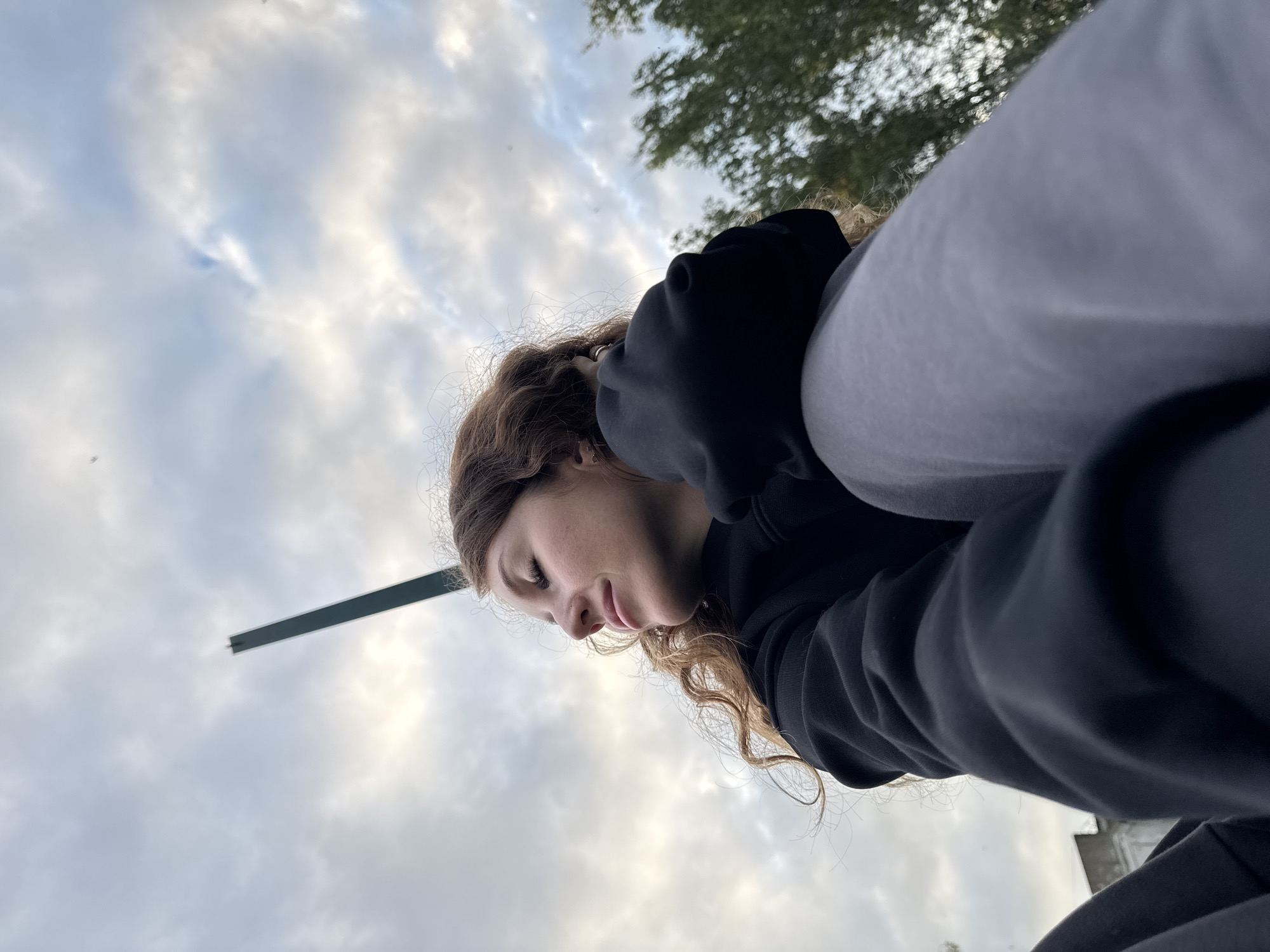 An outdoor, low-angle, medium shot captures a young woman with wavy brown hair looking upward and to the left, her eyes closed and a slight smile on her face. She is wearing a black hooded sweatshirt and grey sweatpants. Her left arm is bent, with her hand near her head, and her right arm is bent across her lap. The background consists of a cloudy sky with a thin, dark, vertical object protruding from the bottom left of the frame towards the top center. To the right of the frame, out of focus, is a cluster of green trees and a blurry building structure. The image appears to have been taken in Chaikovsky, Russia, as indicated by the context.