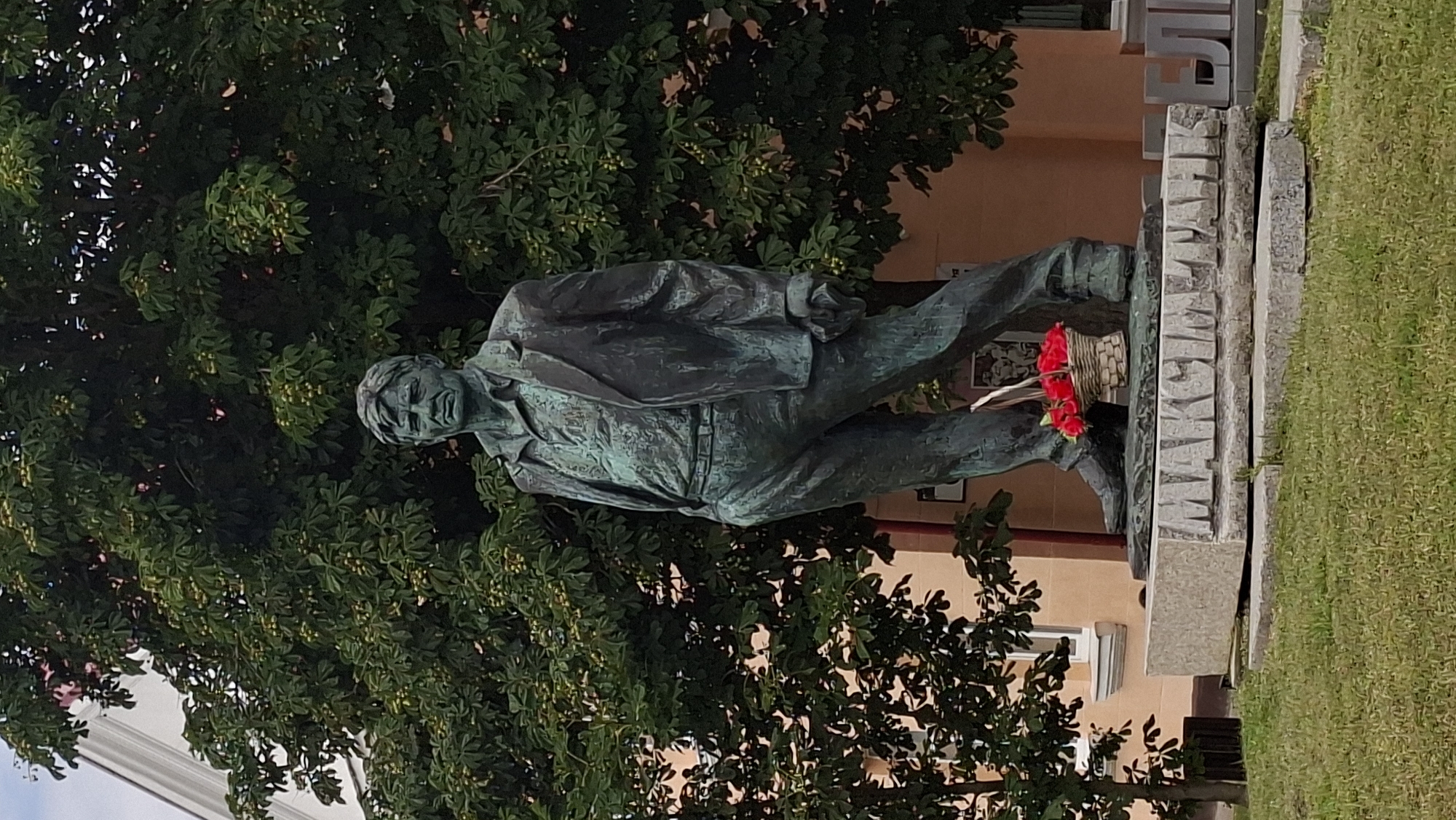 A bronze statue of a man wearing a suit and open-collared shirt stands outdoors, with a basket of red flowers at his feet. The statue is positioned on a grey stone base inscribed with Cyrillic letters that spell "MAKSIM TANK". Behind the statue is a large green tree with visible fruit, and a light orange building with a white-trimmed window can be seen to the left. The statue is located in the city of Myadzyel, Belarus. The ground in front of the base is covered in green grass.