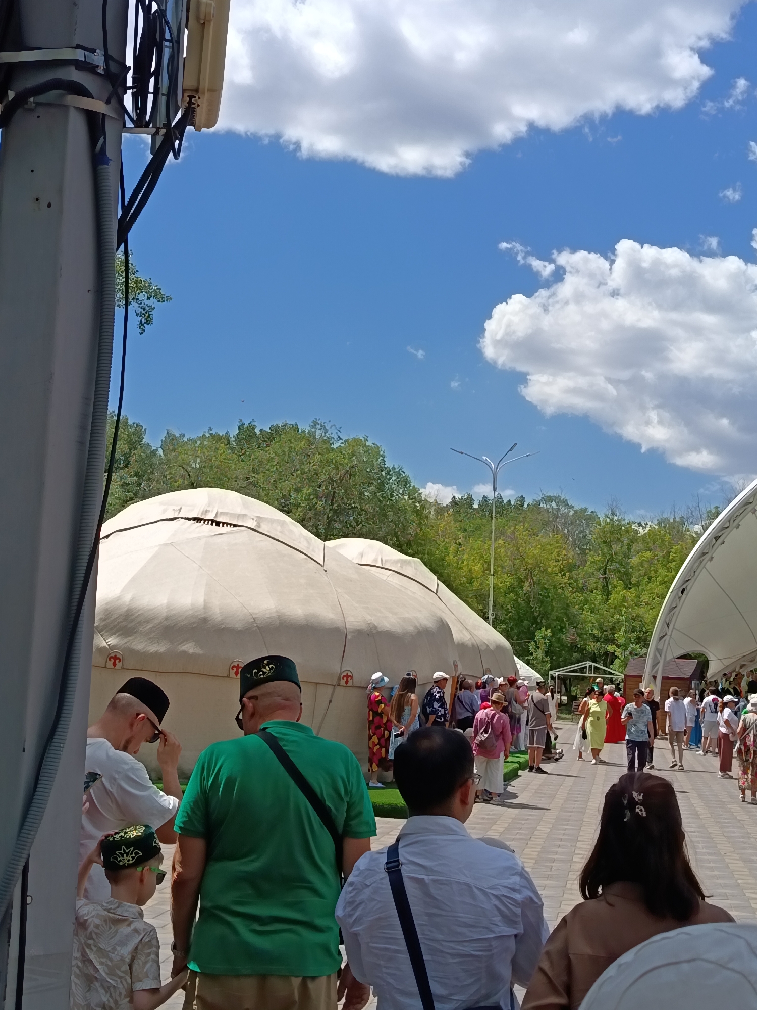 The image depicts an outdoor event with a crowd of people near large, yurt-like structures and a white tent. In the foreground, several individuals are visible, including men wearing traditional skullcaps and a child in a patterned shirt. The background features lush green trees under a bright blue sky with fluffy white clouds. The ground is paved with light-colored bricks. The scene suggests a cultural festival or gathering in an urban park setting.
